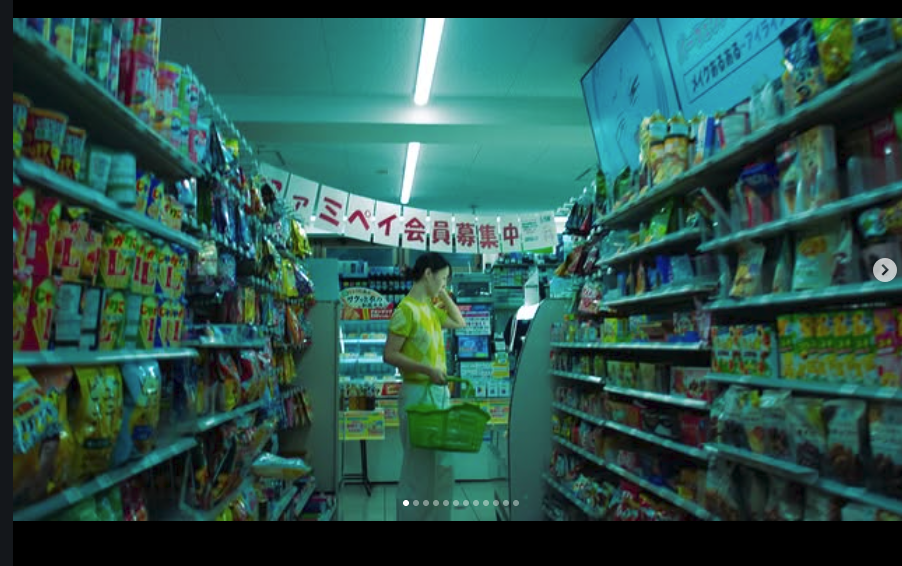 A woman shopping at a grocery store checkout counter with shelves of snack foods on either side and a Japanese sign overhead.
