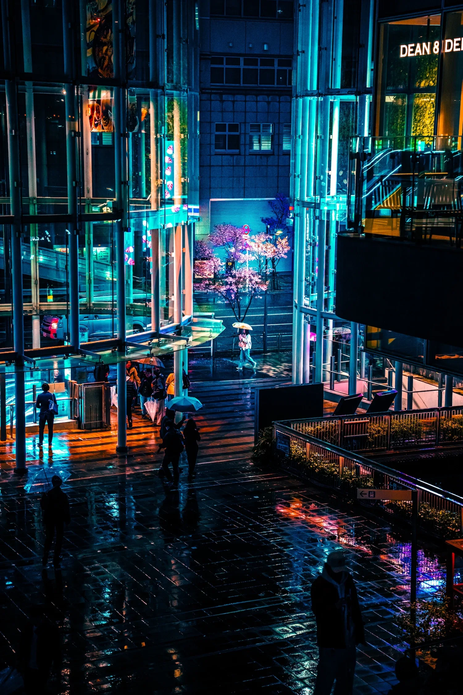 Night scene of a modern city street with glass buildings illuminated by colorful neon lights, a group of people holding umbrellas walking on a wet sidewalk, and some characters inside the buildings visible through the windows.