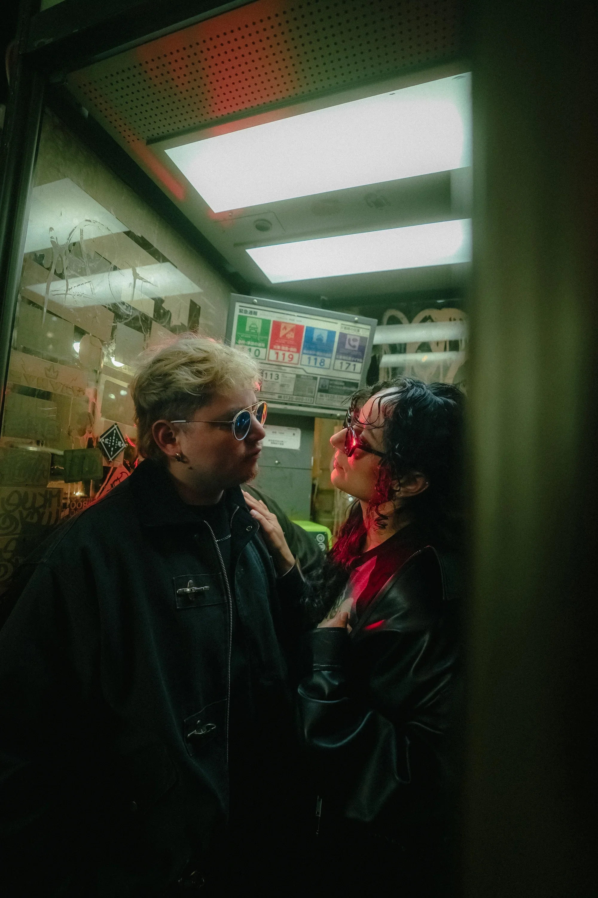 Two people, one with blonde hair and glasses and the other with dark curly hair and red glasses, are facing each other in a confined space, possibly inside an elevator, with a digital screen displaying bus or subway information behind them.