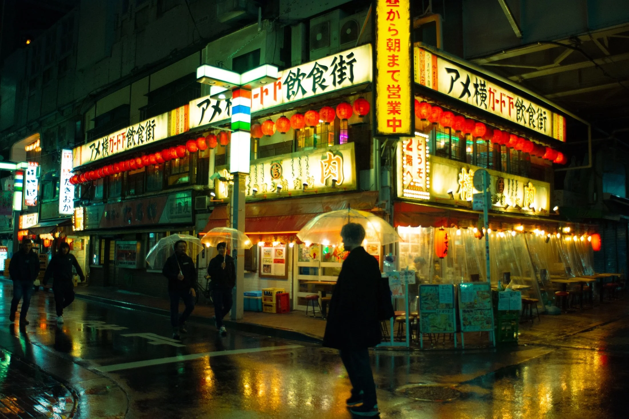 Nighttime street scene with illuminated signs and red paper lanterns, people walking with umbrellas on a wet road.