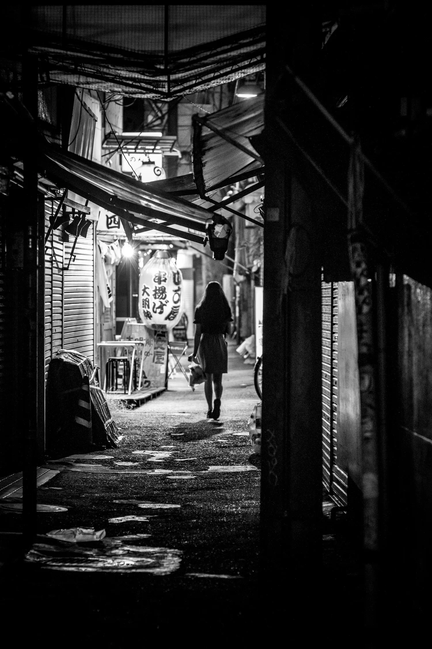 A person walking through a deserted alley at night, lined with closed shops and illuminated by a lantern hanging from a shop entrance.