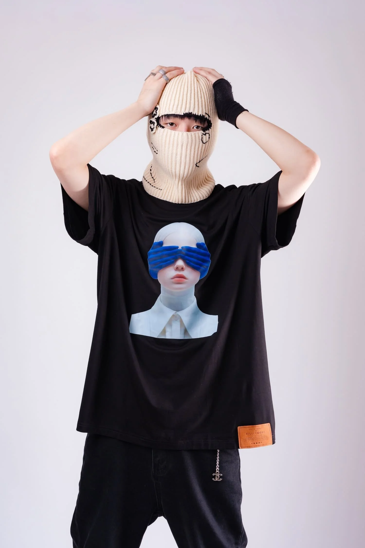 Fashion studio portrait of a model wearing a beige horned knit balaclava mask and oversized graphic t-shirt featuring a surreal portrait with blue hands covering the eyes, photographed by Tokyo photographer Deniz Demir.