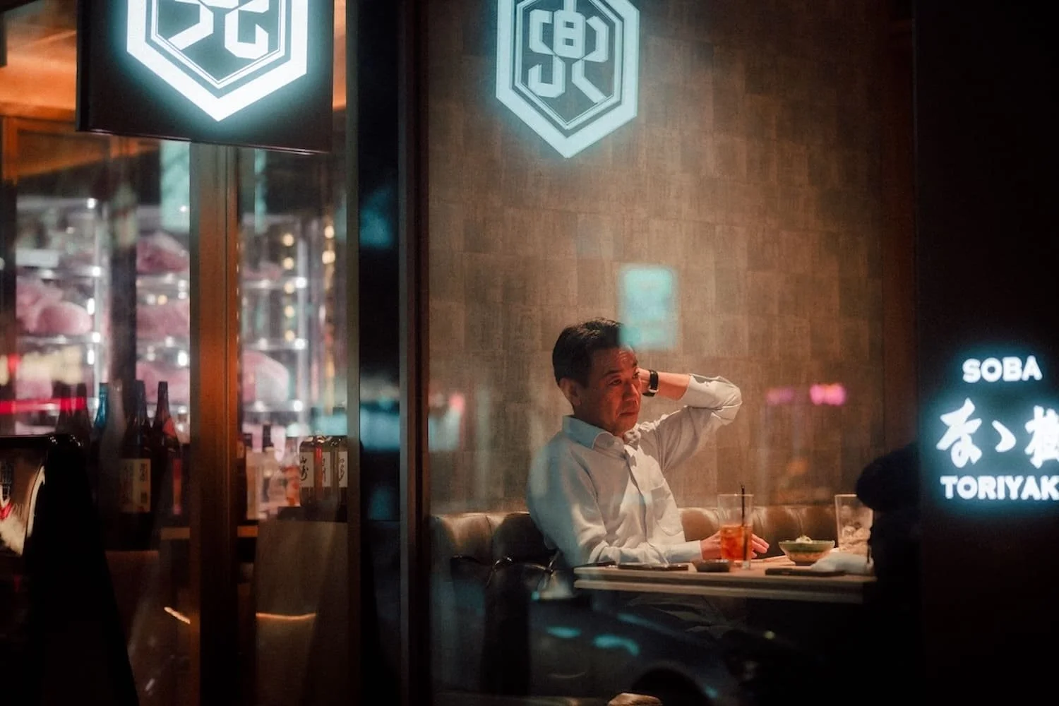 A man sitting inside a restaurant at a table, dressed in a white shirt, with a glass of drink and food on the table, seen through a glass window with reflections and signage.