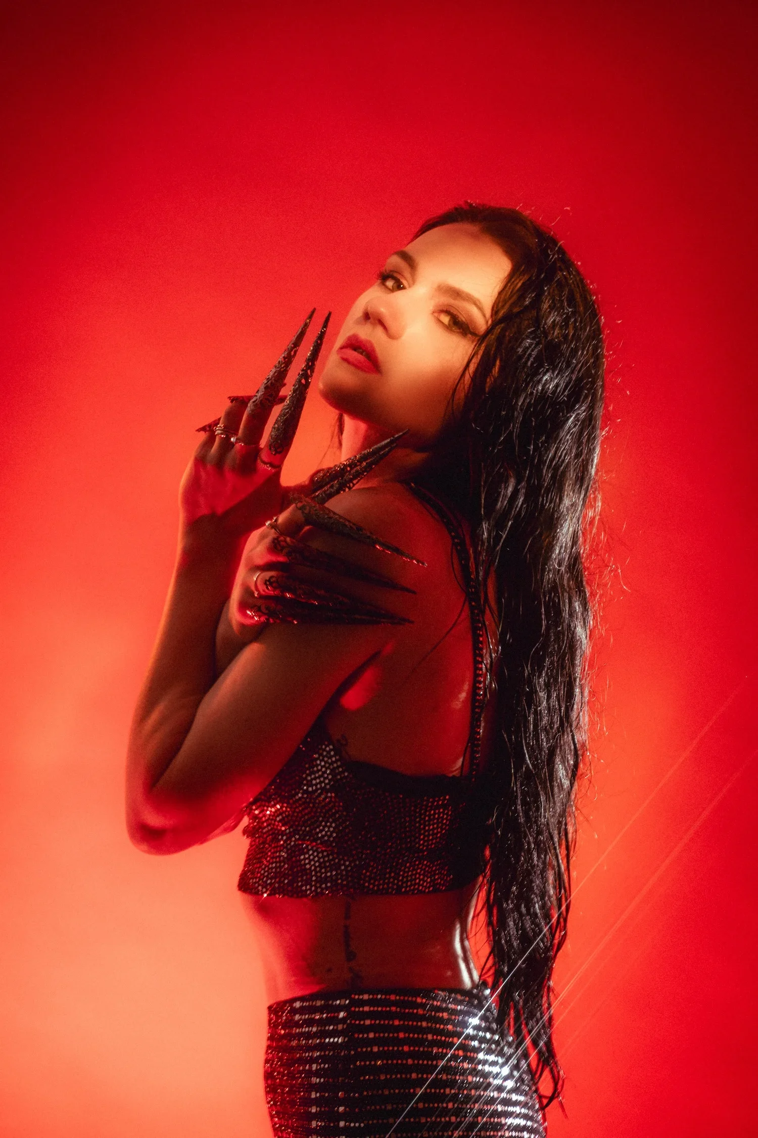 A woman with long dark hair and makeup, wearing a shiny, black, semi-transparent outfit with intricate designs, against a red background with dramatic lighting.