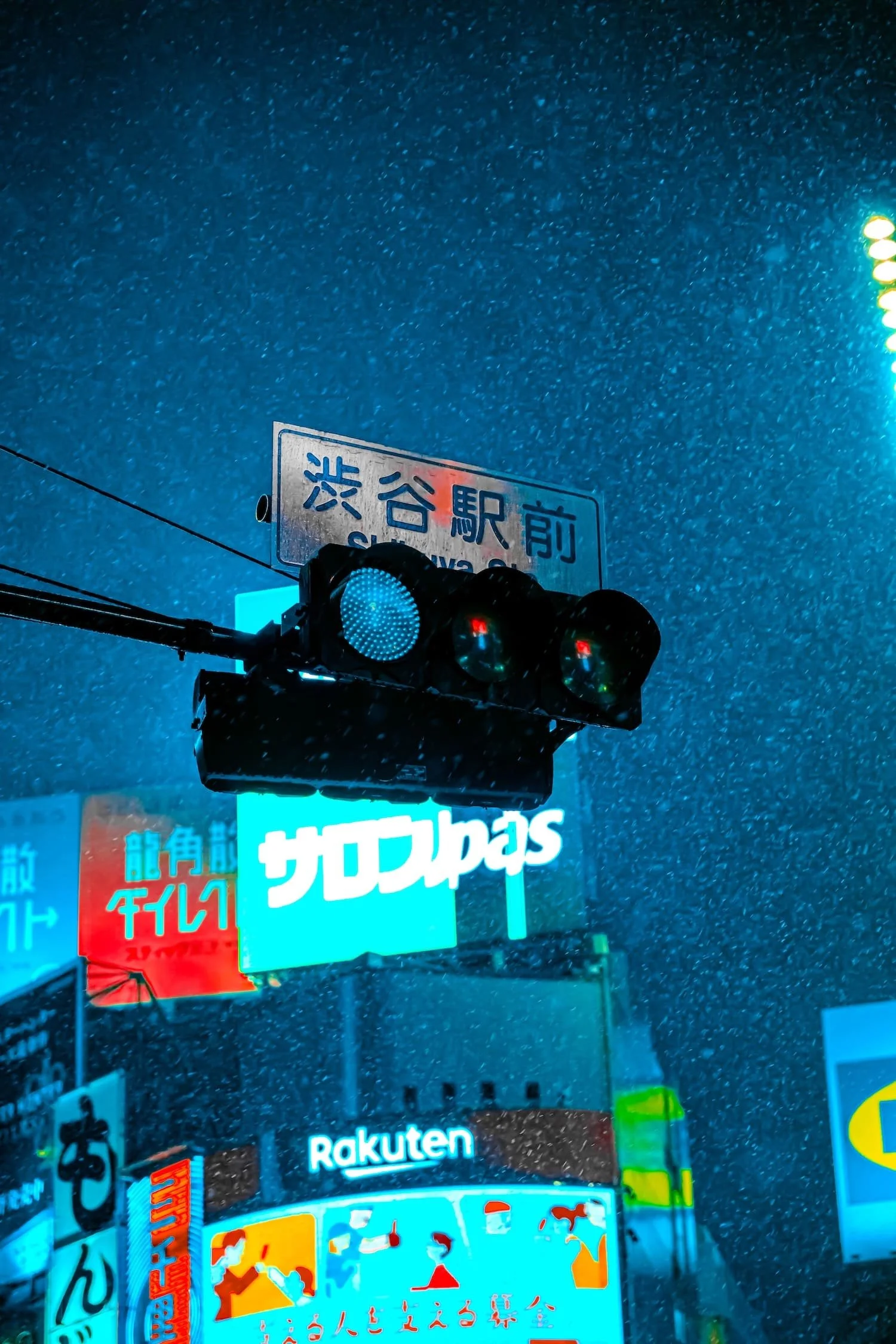 A rain-soaked city street at night featuring illuminated neon signs in Japanese characters, a traffic light with red signals, and a billboard for Rakuten.