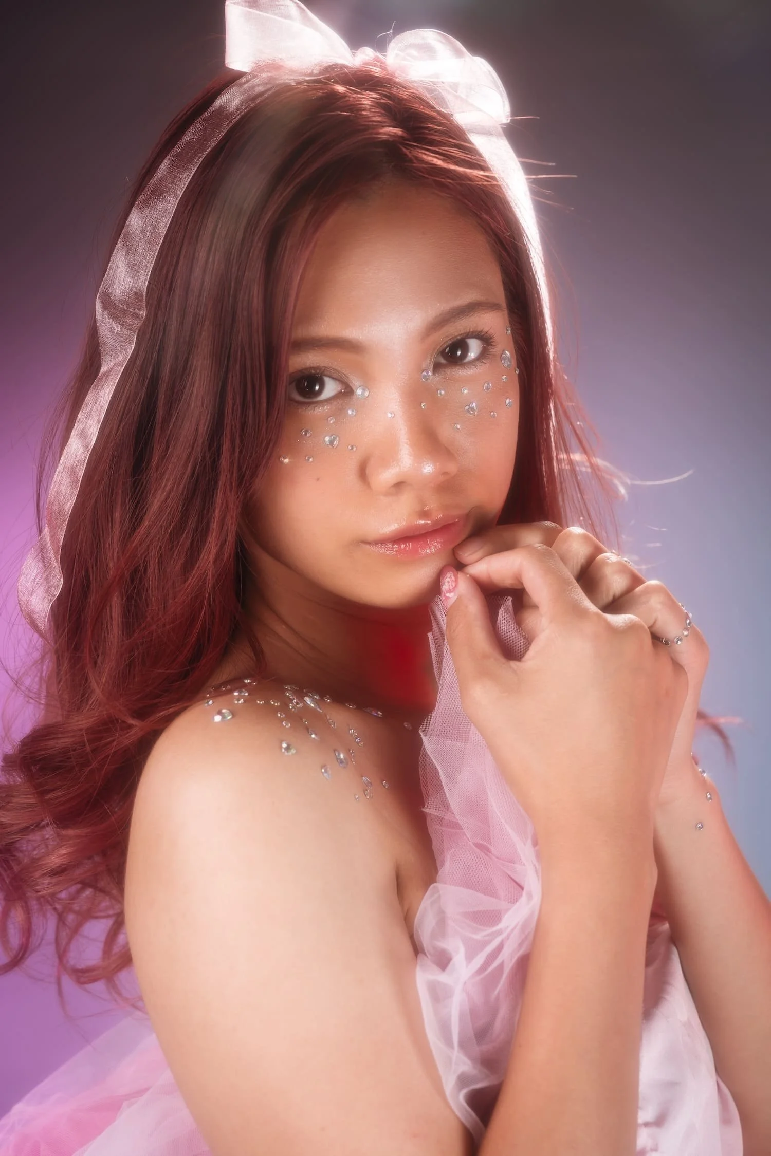 A young woman with auburn hair styled in loose waves, wearing light pink tulle fabric and decorated with rhinestones on her face, shoulder, and arm. She has a large pink bow in her hair and a dreamy, soft expression.