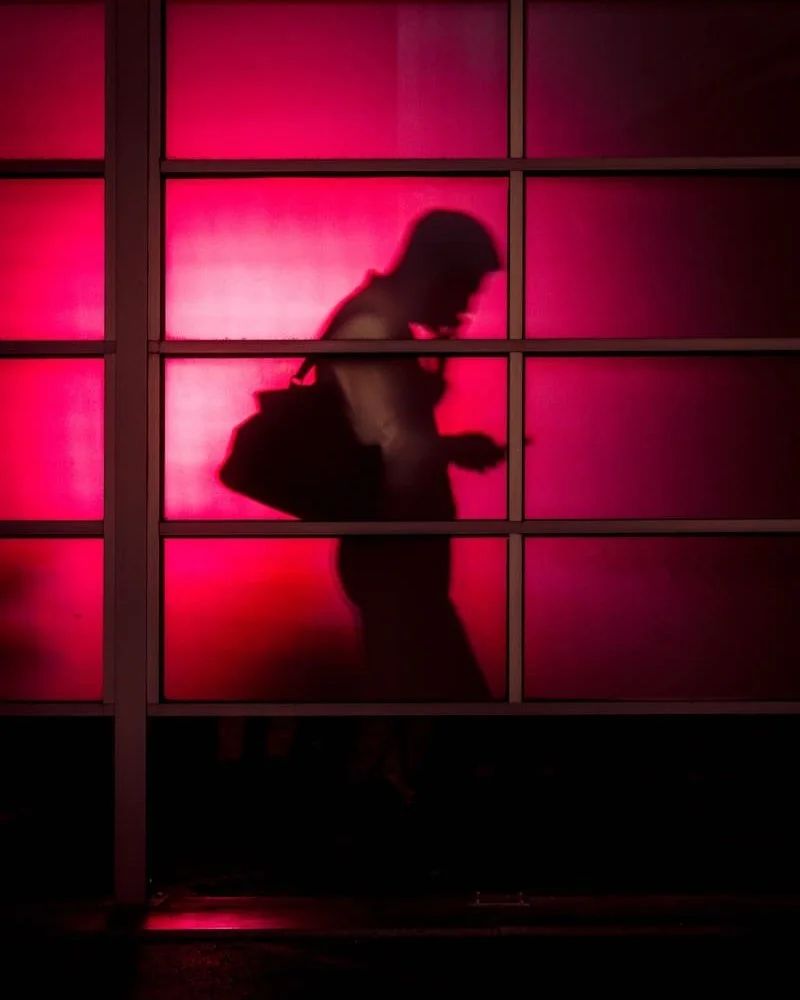 Silhouette of a person with a backpack looking at their phone, seen through a window with pink lighting.