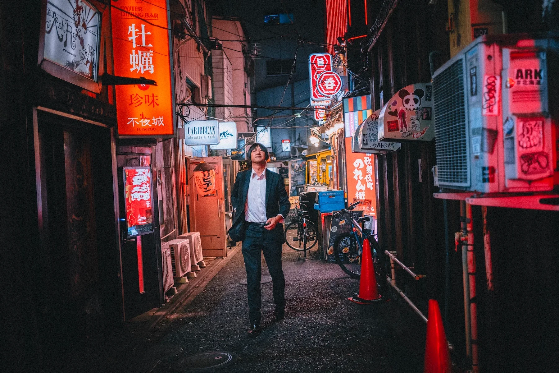 A man in a suit walking on a narrow, nightlife street in Japan, illuminated by neon signs and colorful lights, with bicycles and various small shops or bars lining the street.
