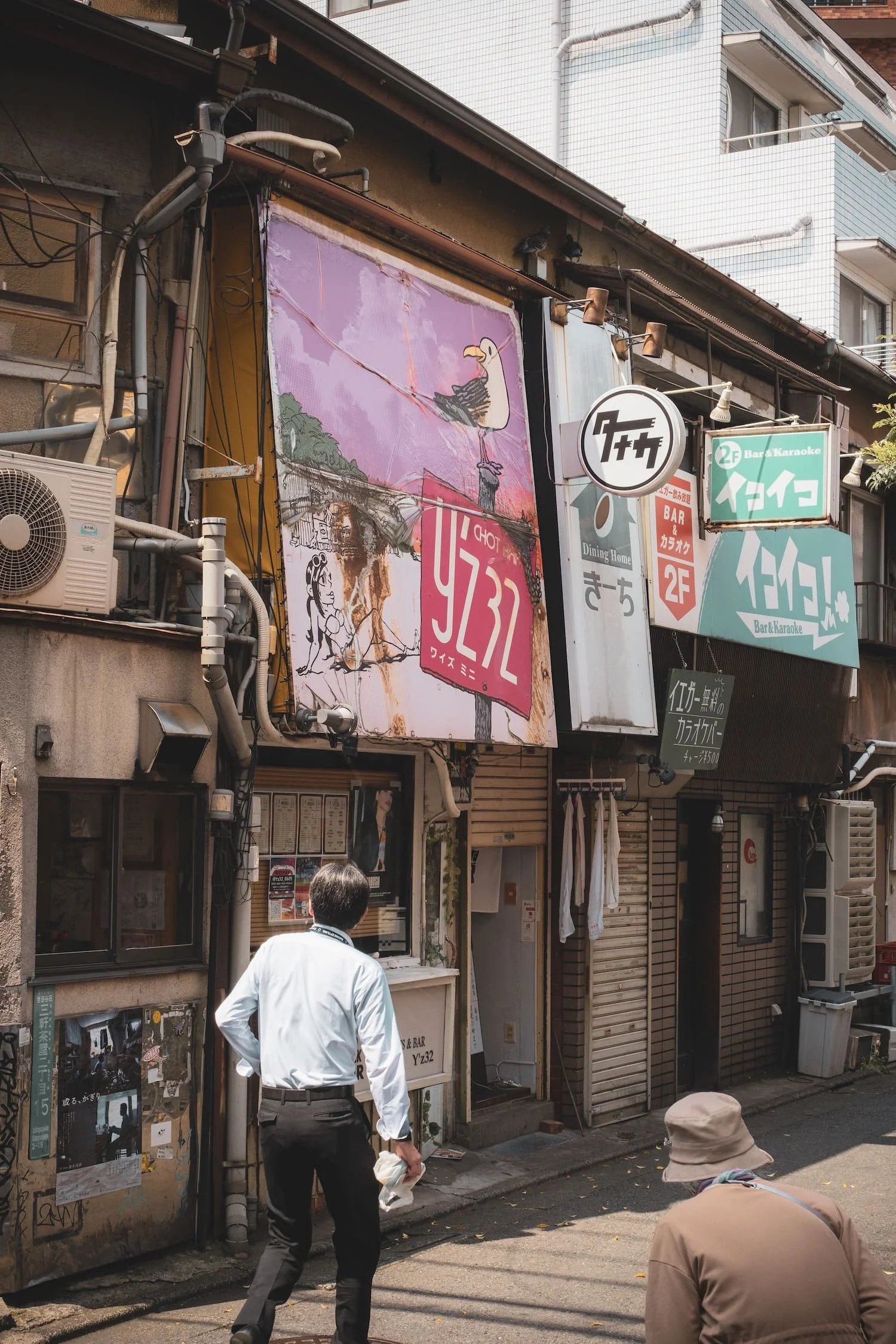 Street view of a building with various signs and a large advertising banner, two people walking in the foreground.