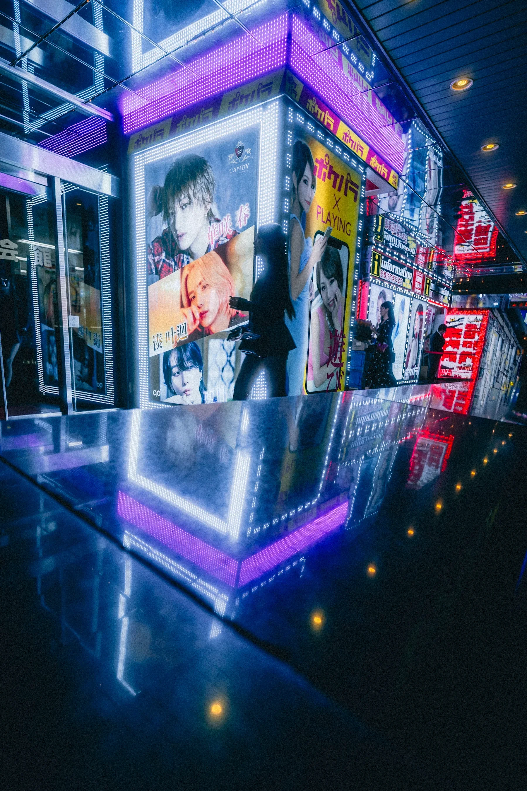 Brightly lit Digital Advertising panel with images of young women, neon lights, reflections on the floor, and various signs in a busy urban area at night.