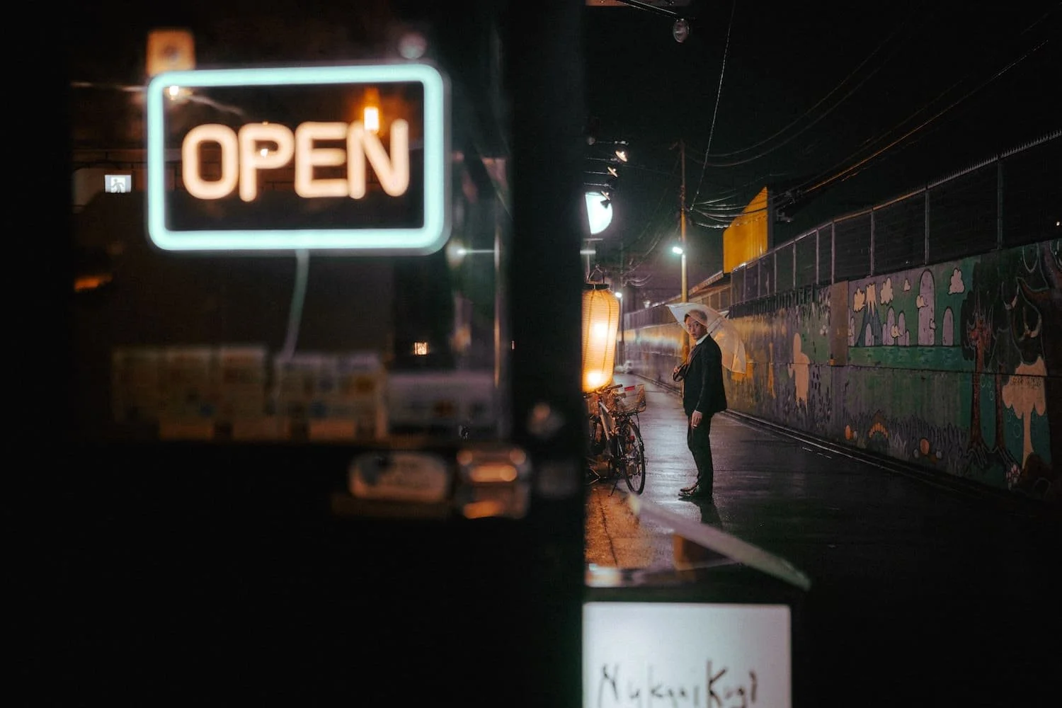 A man holding an umbrella waits on a rain-soaked street at night, illuminated by streetlights and colorful murals on a wall behind him, with a neon "OPEN" sign in the foreground.
