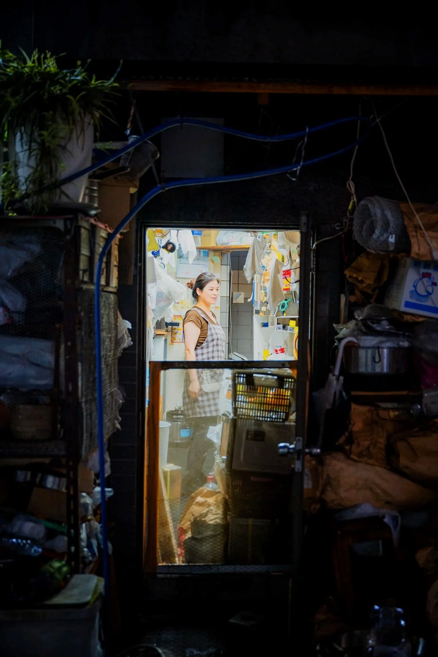 A woman standing inside a small, cluttered shop or kitchen, smiling, with various items hanging and stored around her, illuminated by indoor lighting, with a dark exterior seen through a doorway.