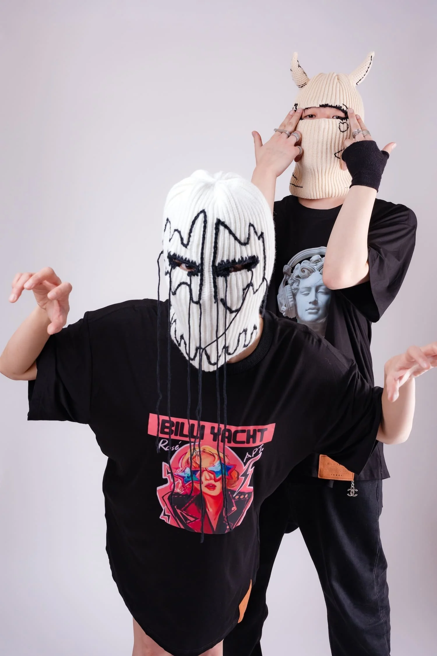 Studio fashion portrait of two masked models wearing oversized graphic streetwear t-shirts and knit balaclava masks, posing playfully against a minimalist white background, photographed by Tokyo photographer Deniz Demir.