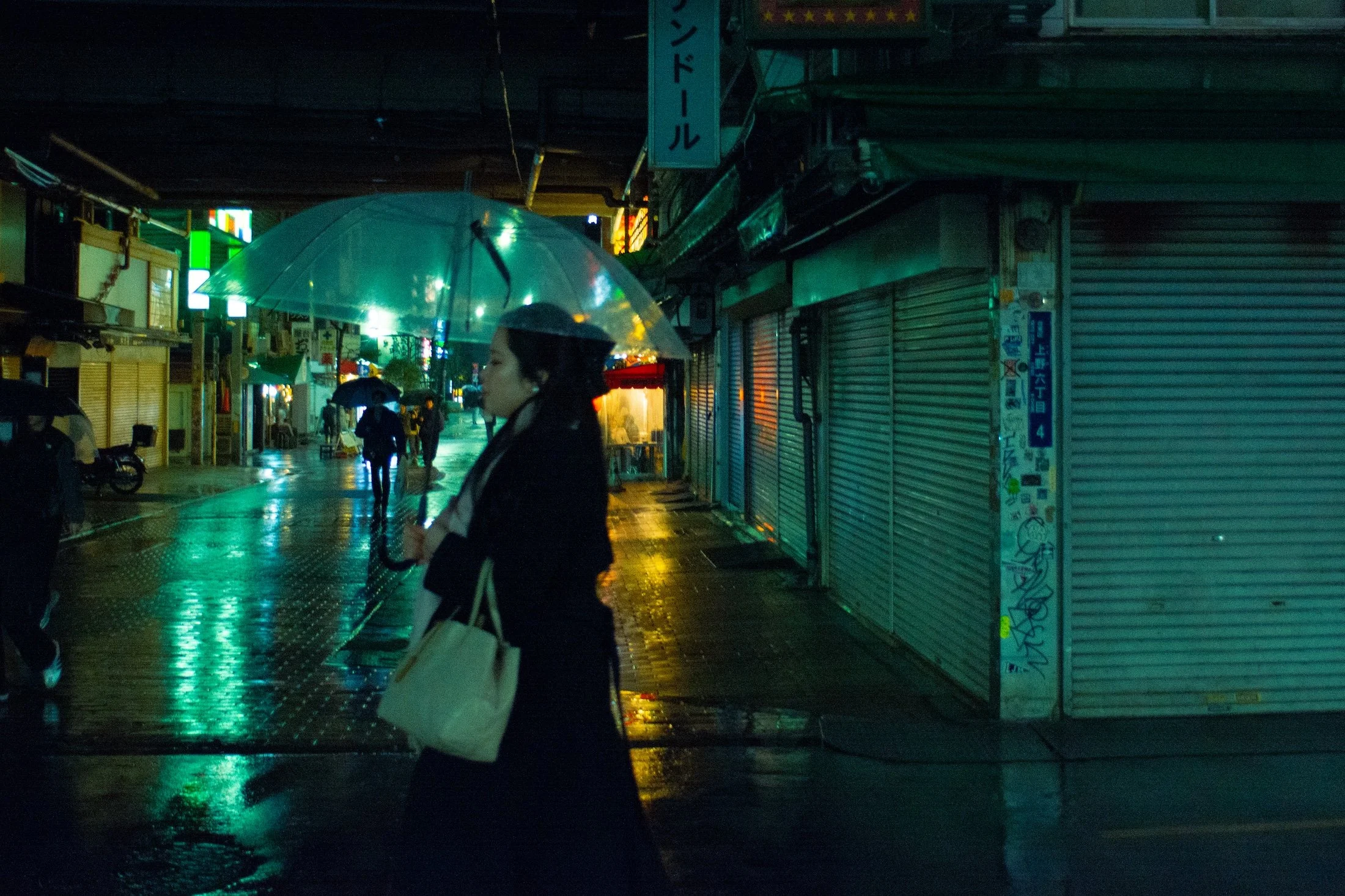 A woman walking with an umbrella in the rain on a city street at night with closed shops and neon signs shot in Tokyo at night.