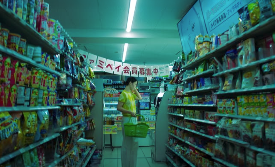 Woman shopping in a convenience store aisle filled with snack foods and beverages, with a Japanese sign overhead.