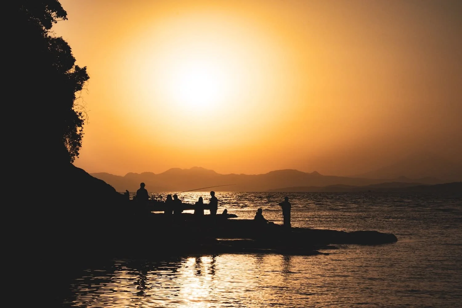Silhouettes of people fishing and sitting on rocks along a shoreline at sunset, with mountains in the background and the sky glowing orange. Tekumi photography workshop called Shoreline Stories.
