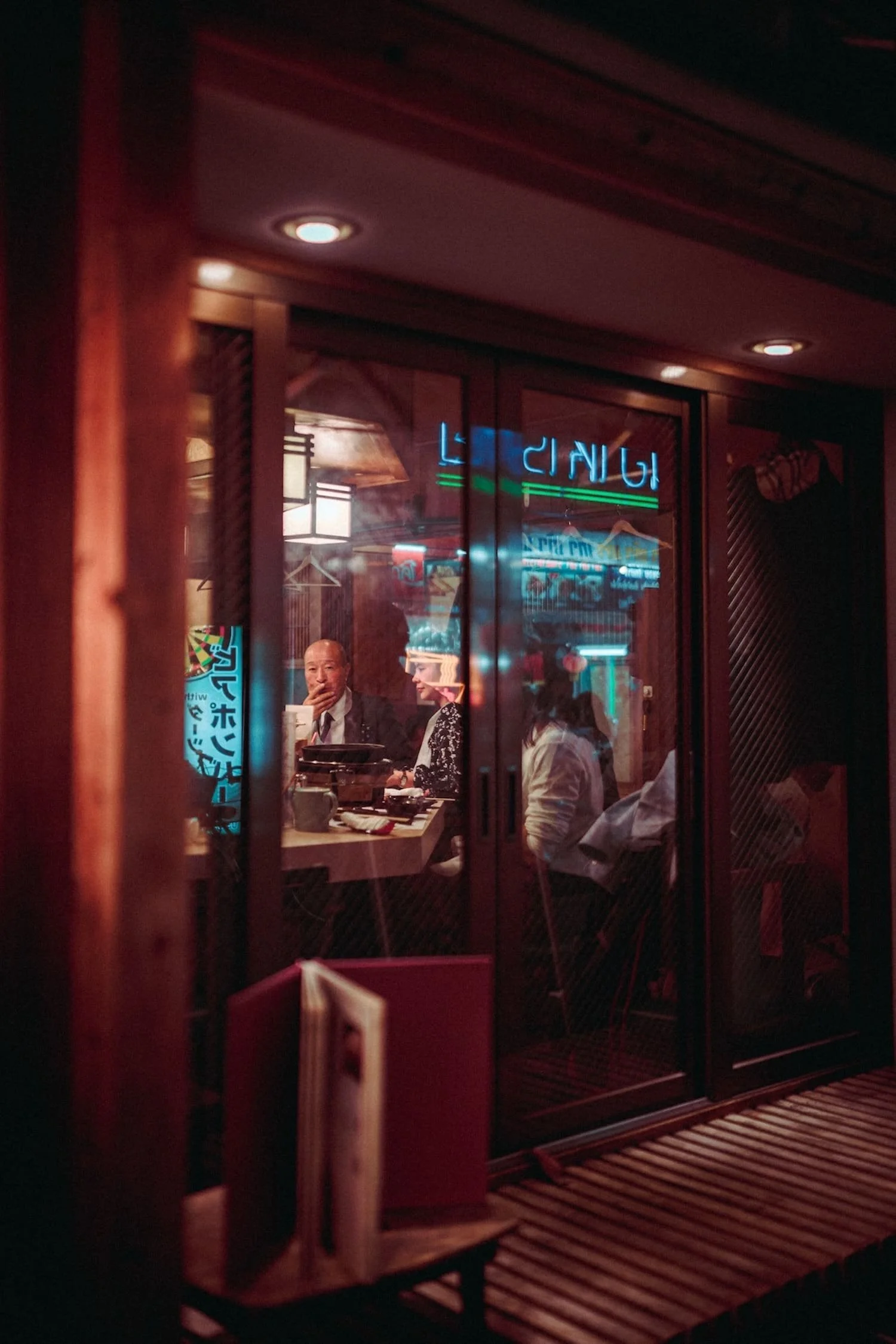 People dining inside a restaurant seen through glass doors with neon signs reflecting on the glass.