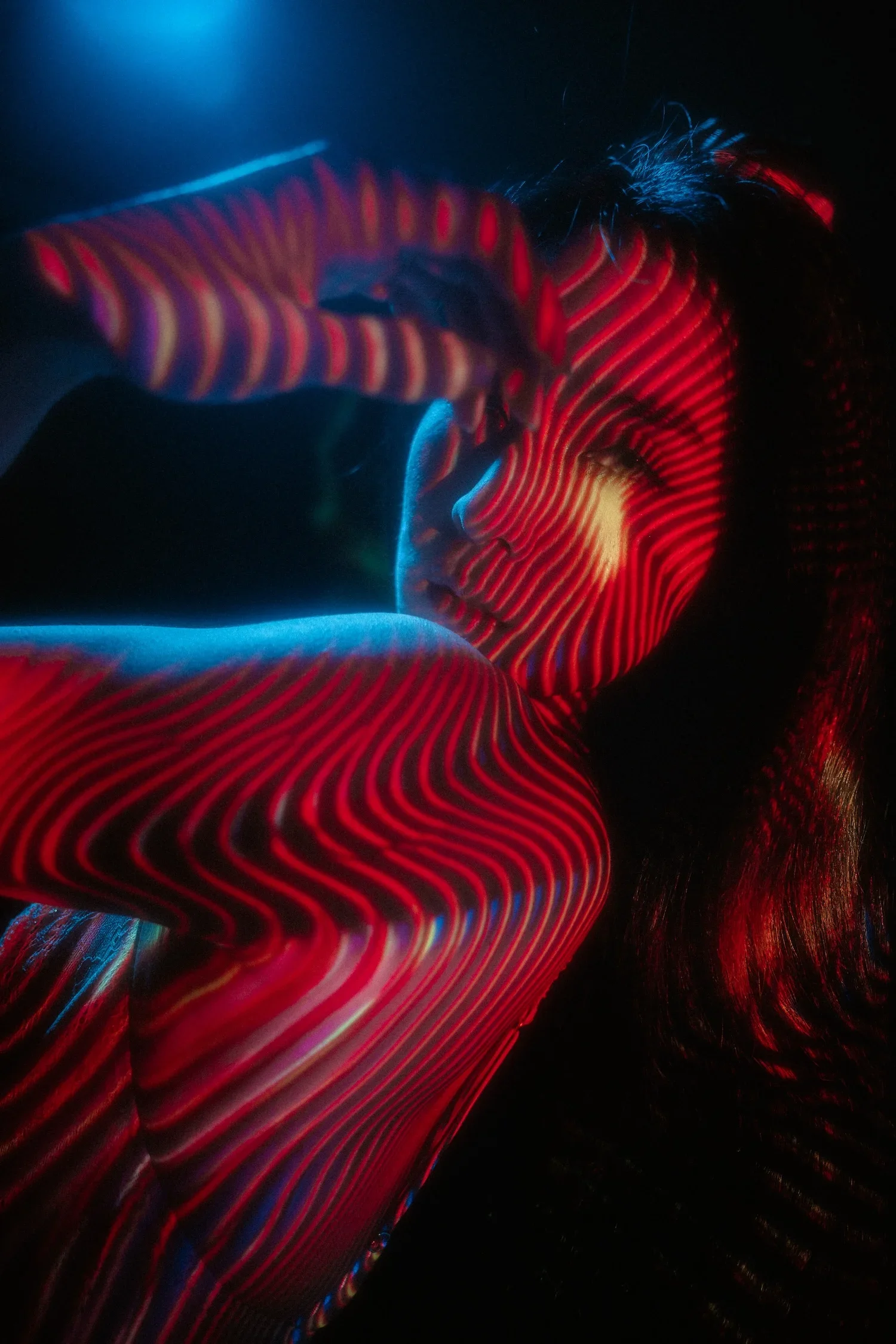A person with curly hair is lying down in the dark, illuminated by colorful, wavy light patterns projected onto their face and upper body, creating an abstract, vibrant visual effect.