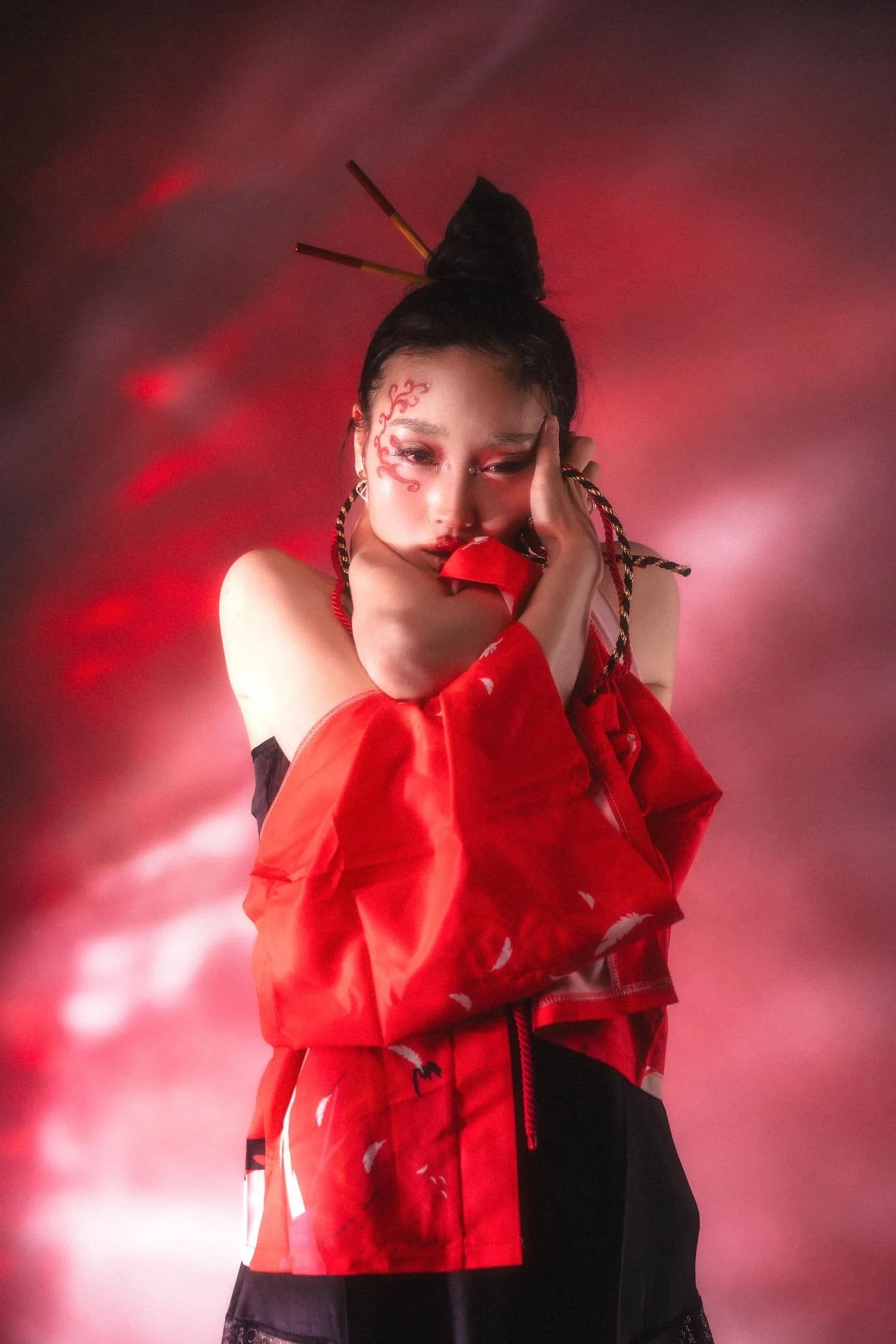 A beautiful Japanese model with facial makeup and hair styled with two chopsticks, wearing a red satin jacket, poses with her hand on her face against a red and black smoky background.