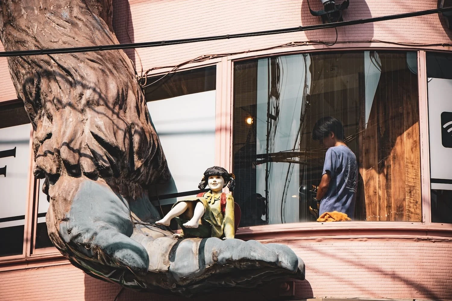 Decorative window feature with a large sculpture of a hand holding a small girl figurine on the outside of a pink building, with a person inside visible through the glass window.