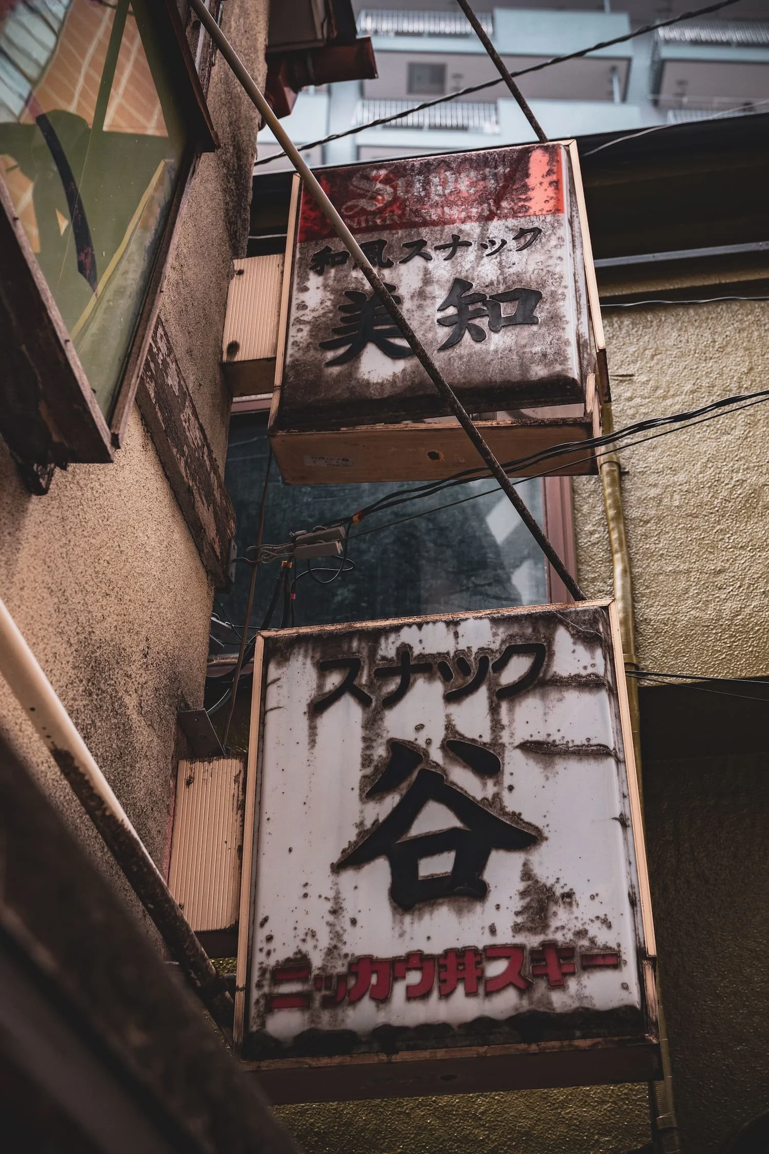 Two weathered, rusted Japanese signs hanging on an exterior wall, with Japanese characters and some black and red text.