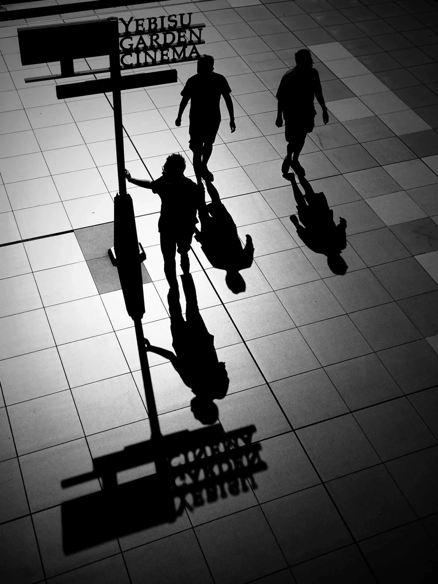 Silhouettes of three people walking on a tiled floor, casting shadows. Signage in the background reads 'YEBISU GARDEN CINEMA'.