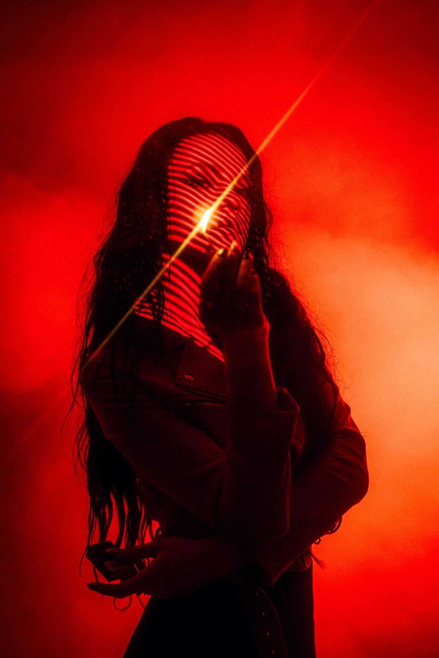 A woman with long, curly hair is illuminated by red and yellow laser light projections against a red background, creating stripe patterns across her face and body shot by Tokyo photographer Deniz Demir.
