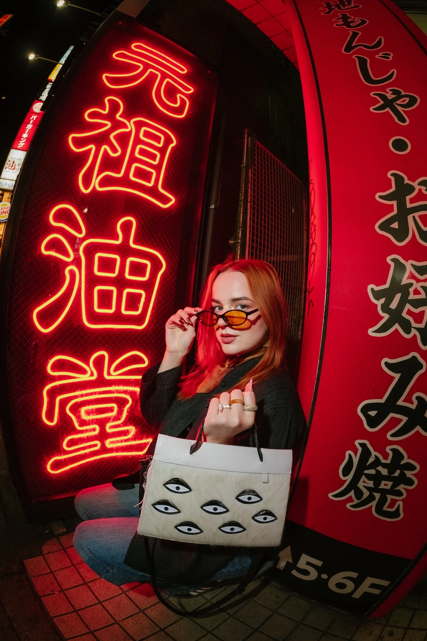 A woman with red hair and sunglasses sitting by a neon-lit Japanese sign on a city street at night. She is holding a shopping bag with eye illustrations.