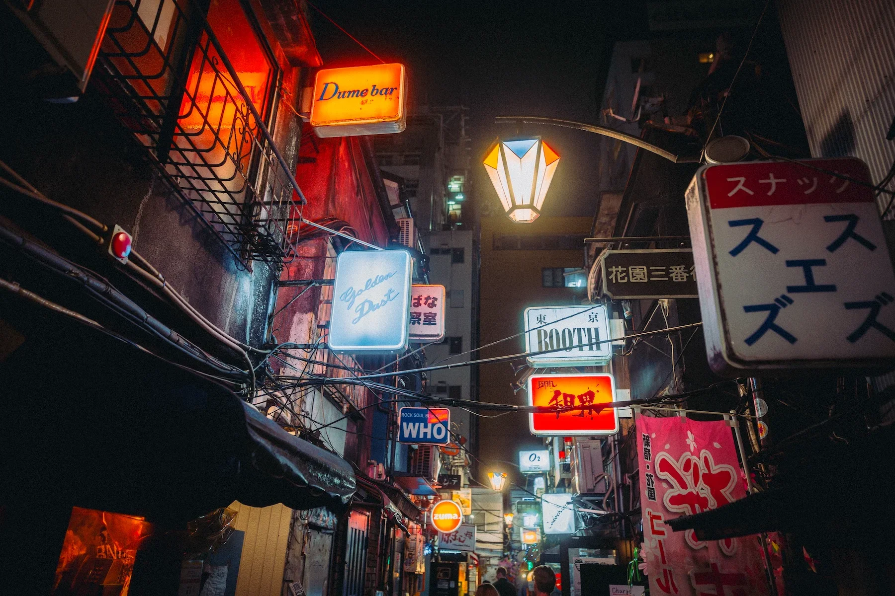 Night scene of a narrow street in Japan illuminated with various colorful neon signs in Japanese and English, some hanging from buildings and others attached to poles.