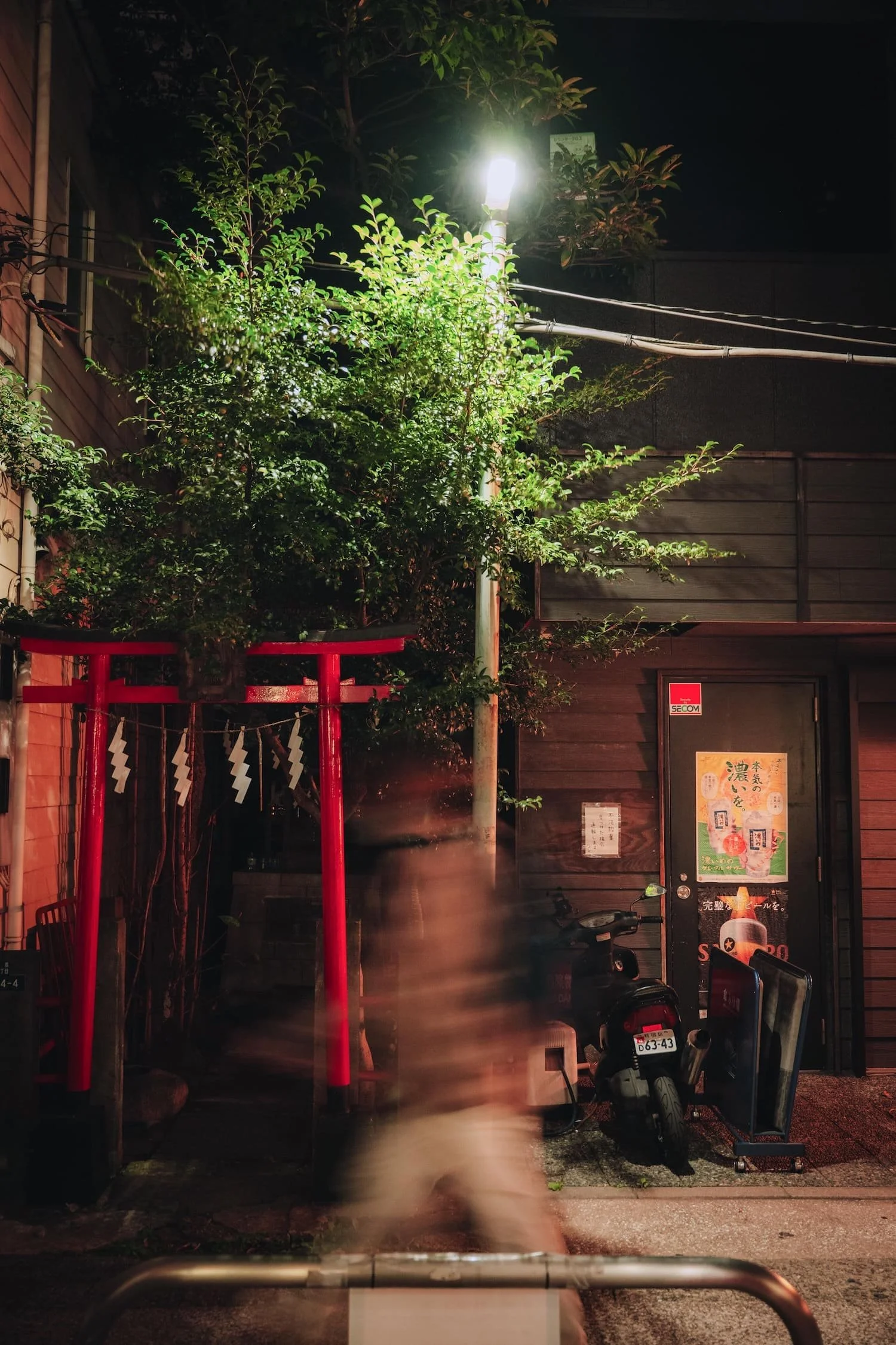 Nighttime scene with a red torii gate, a large green tree, a street lamp, and a black scooter parked near a wooden building with posters on the door. A blurry person is walking in front.