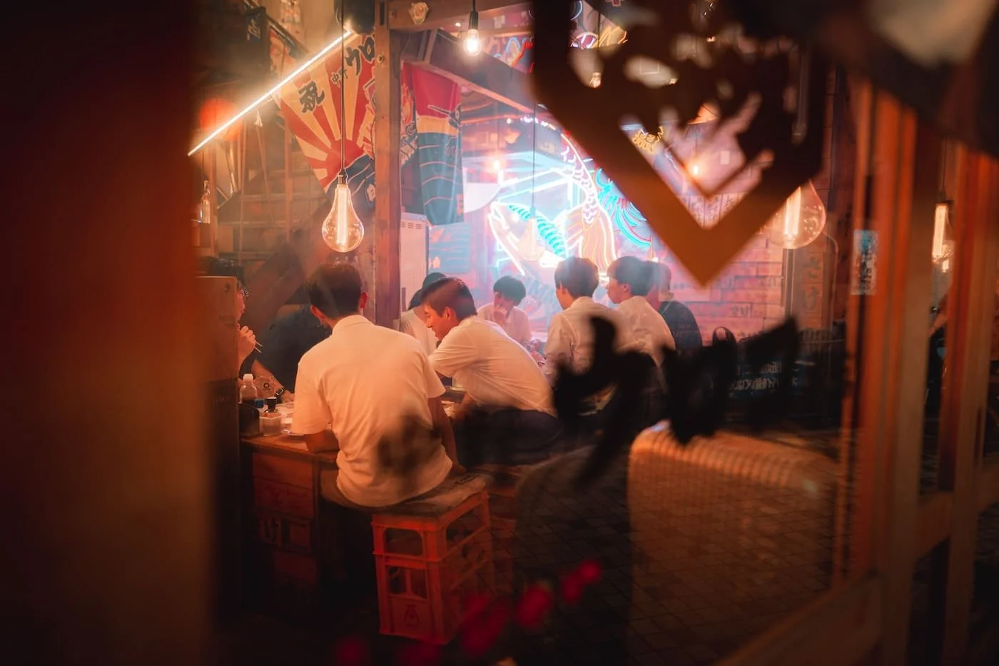A group of young men sitting around a table in a dimly lit Asian restaurant or bar, decorated with neon lights, colorful posters, and hanging light bulbs, viewed through a window with a decorative frame. Tekumi street photography workshop Tokyo.