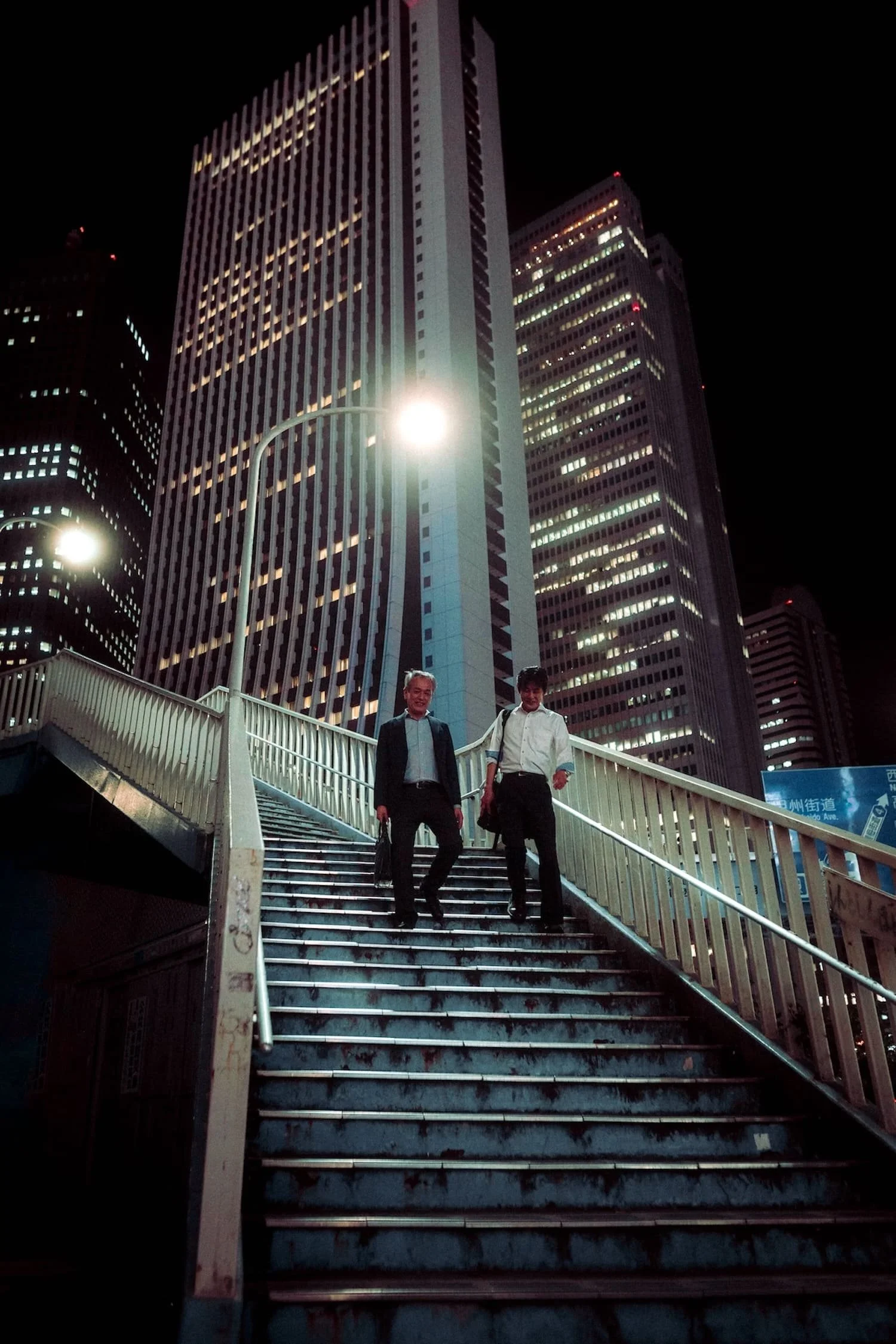 Two businessmen walking down a city stairs at night, illuminated skyscrapers in the background.