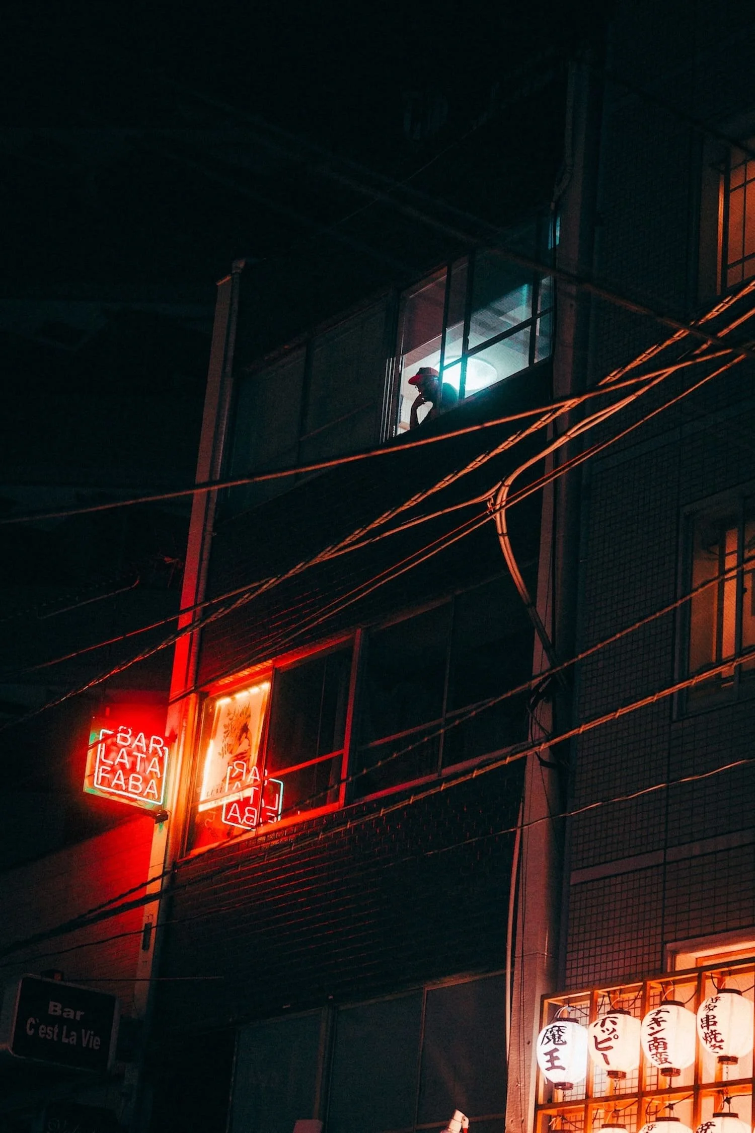 A person leaning on the window of a high-rise building at night, illuminated by blue light. Neon signs in red and lanterns with Japanese characters are also visible against a dark background.