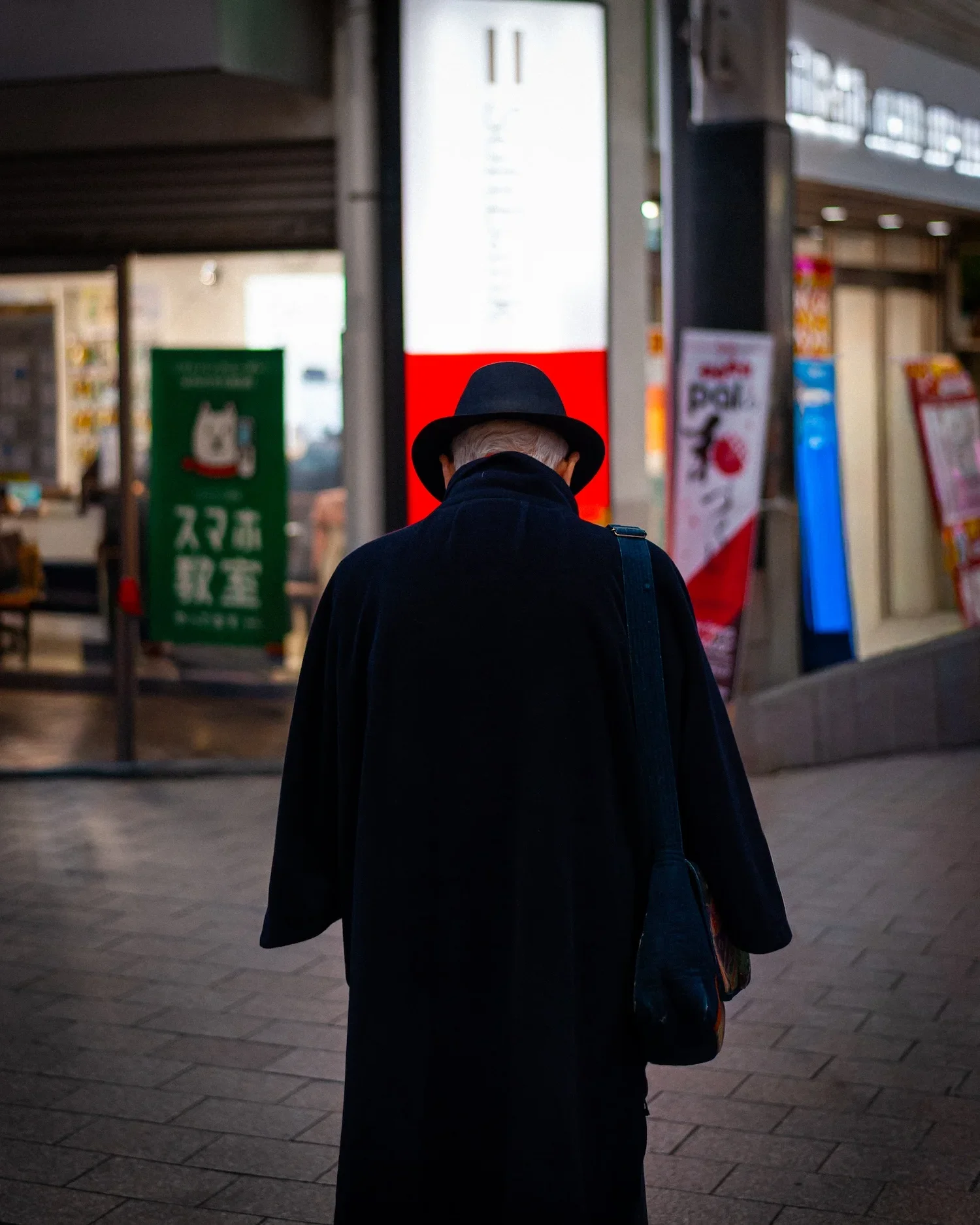 A person wearing a black hat and black coat with their back facing the camera, standing outside near a brightly lit building with signage and advertisements.