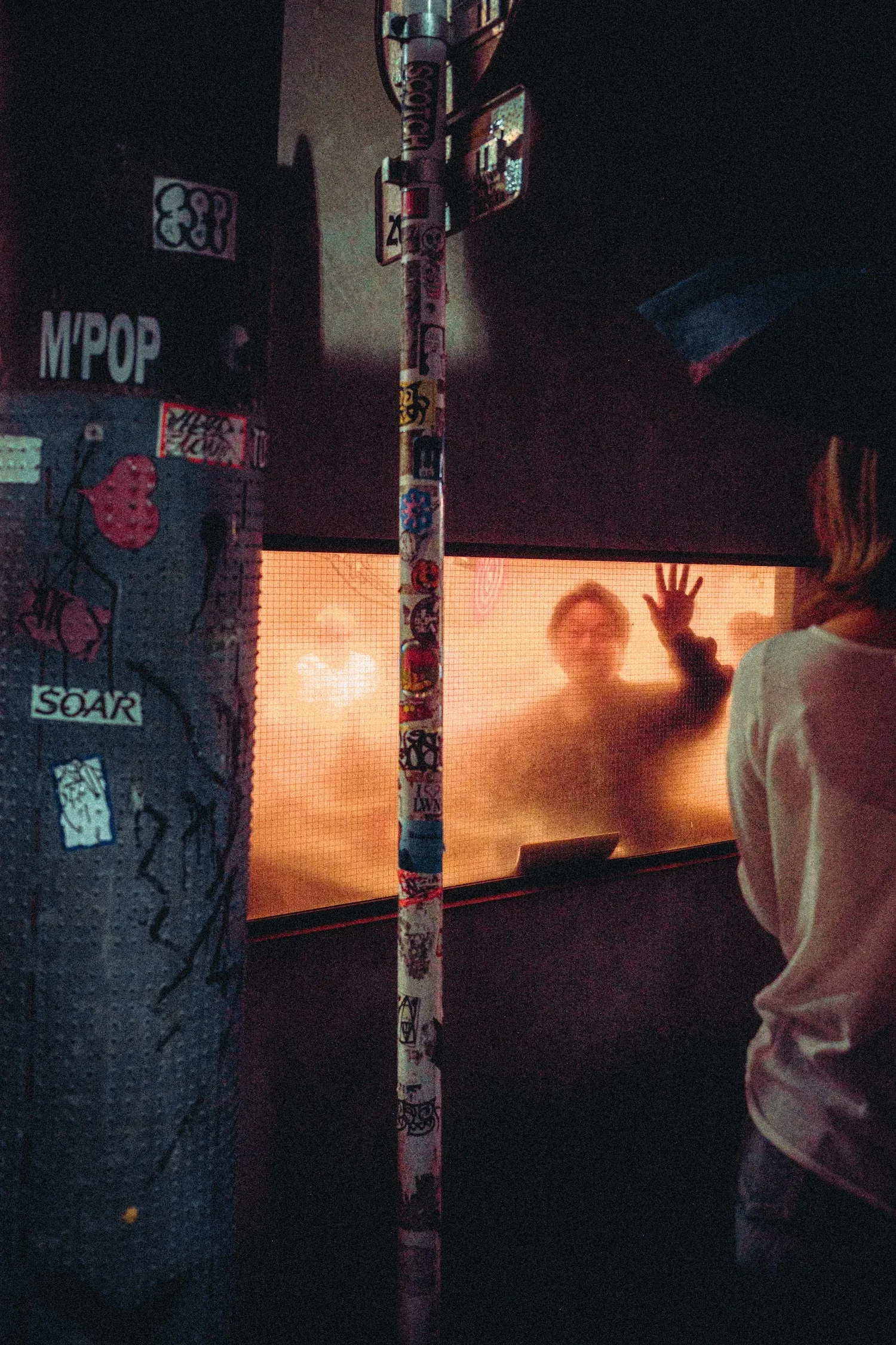 Person standing in front of a foggy, orange-lit window with their hand raised. The scene is viewed through a mesh screen. There are various stickers on a pole and graffiti on the wall nearby.