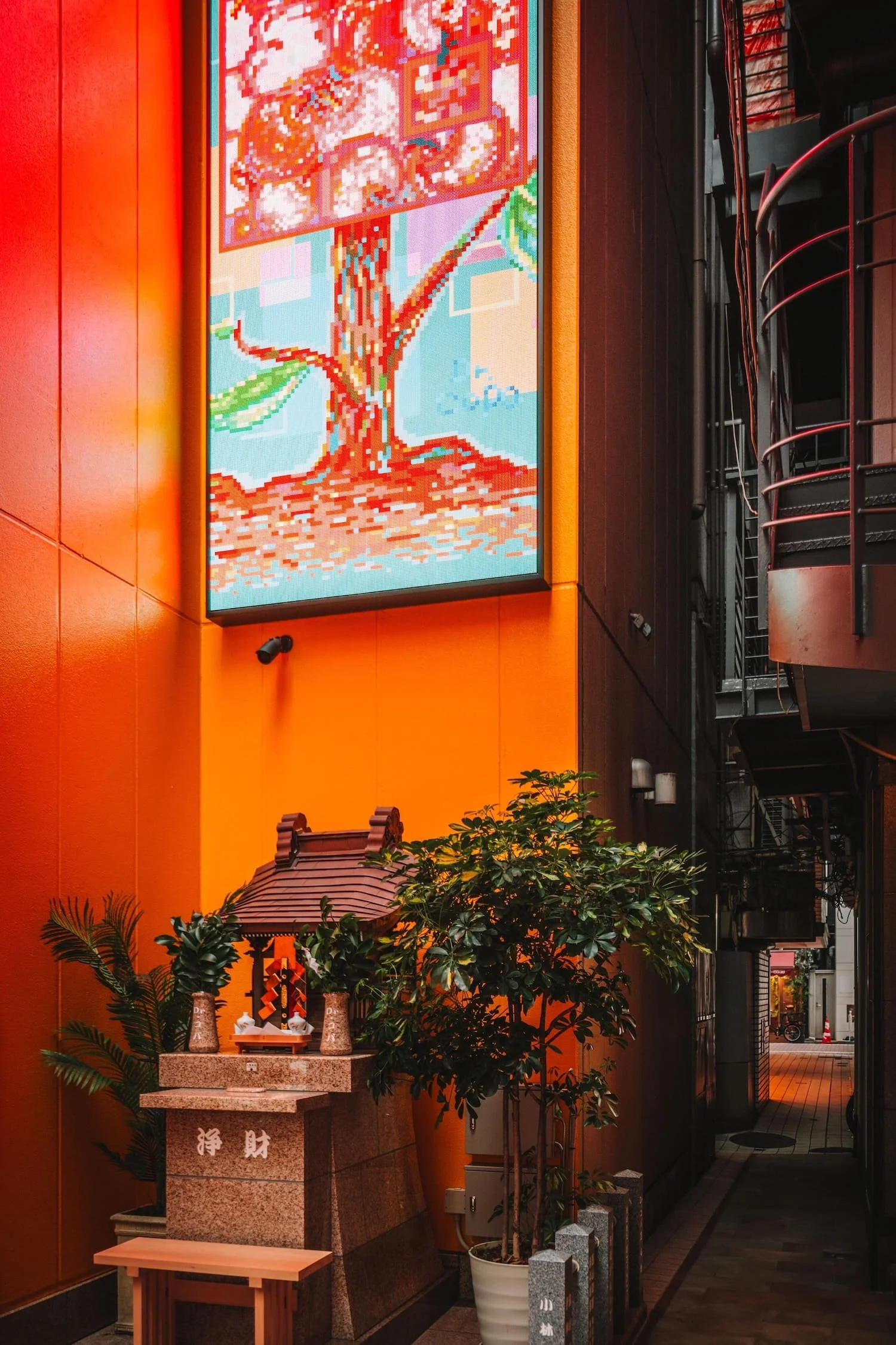 A vertical LED display showing digital artwork of a tree with pixel art style. The display is mounted on a bright orange wall beside a narrow alley. Below the display, there is a small shrine with plants and stone sculptures.