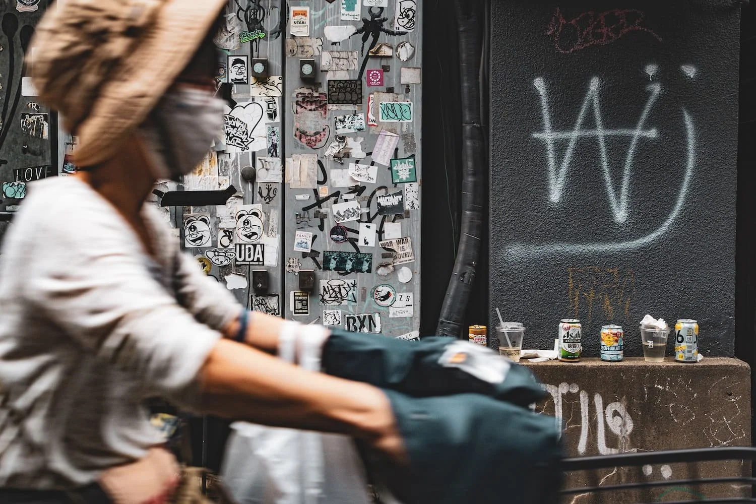 A person wearing a face mask and a straw hat riding a bicycle past a wall covered in stickers, with a black wall on the right featuring a large white graffiti symbol and a table with empty cans and cups.