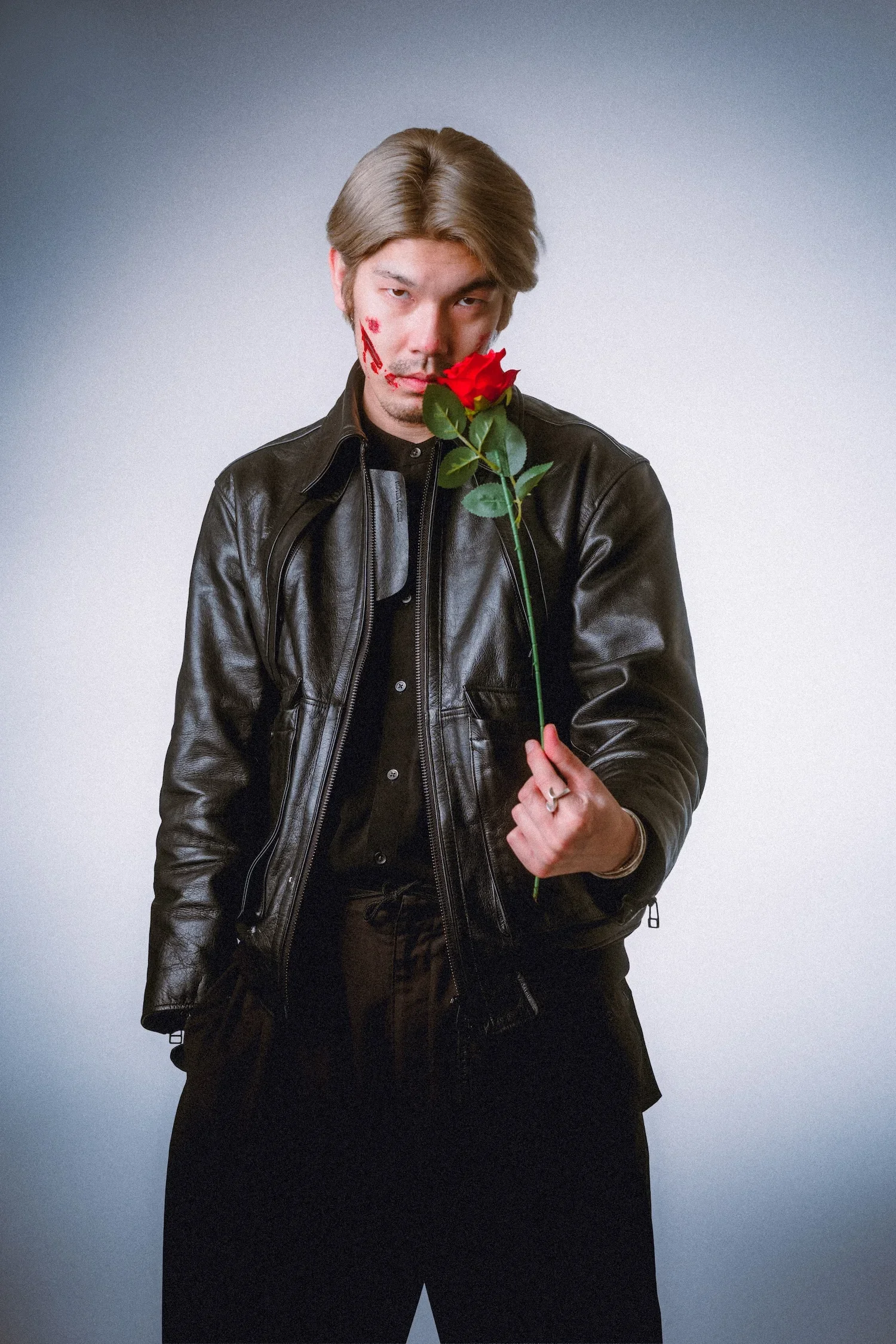 A man with shoulder-length light brown hair holding a red rose in front of his face. He is wearing a black leather jacket and black clothing, standing against a plain gradient background.