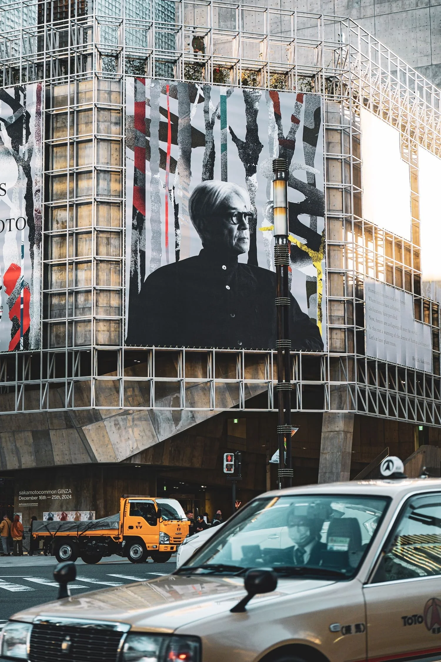 Large building in an urban area with a digital billboard displaying a black and white portrait of a man with glasses. Cars and a small yellow truck are on the street below, with pedestrians on the sidewalk.