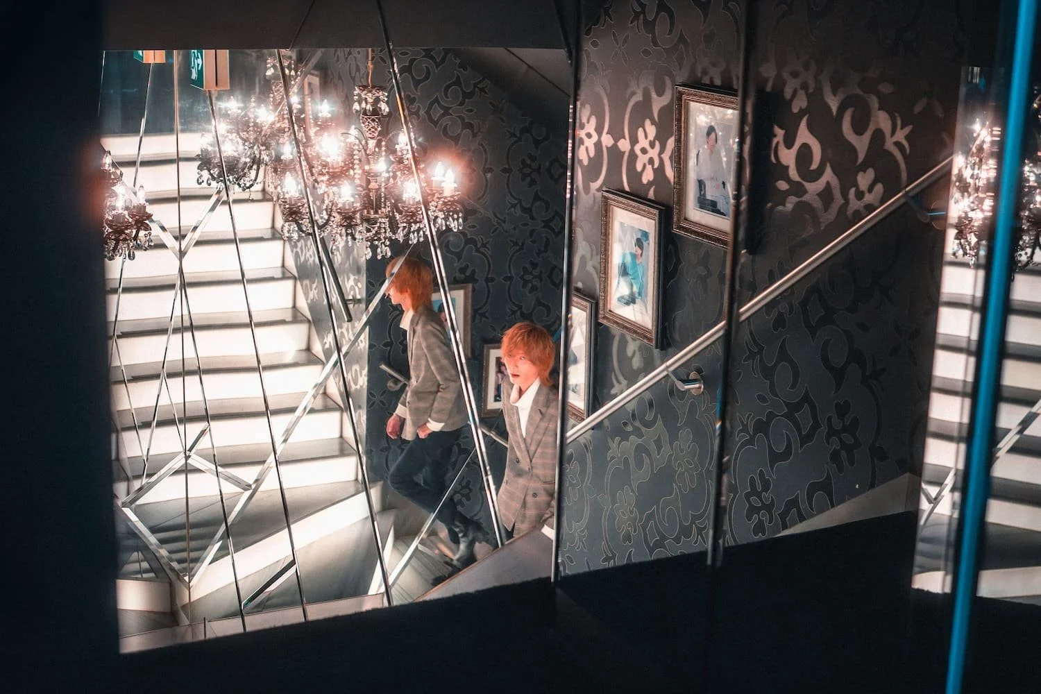A young man with red hair, wearing a checkered blazer and white shirt, walking down a staircase with mirrored walls and framed photographs. The staircase is illuminated with chandelier lights, and the mirror creates a reflective, symmetrical effect.