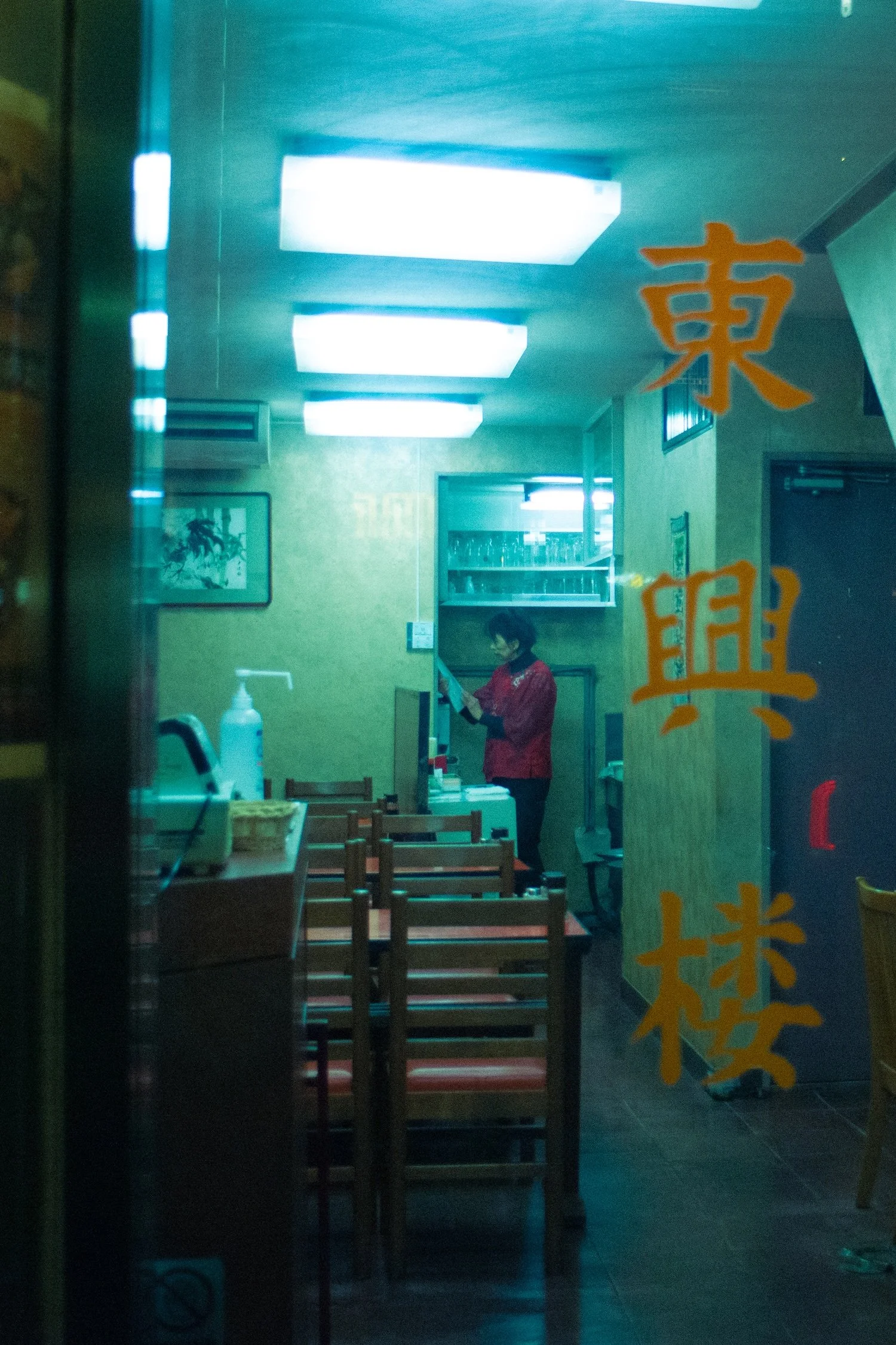 View through a window of a restaurant interior with wooden tables and chairs, a woman in a red jacket standing inside, bright fluorescent ceiling lights, and Asian-inspired artwork on the walls. The window has orange Chinese characters written on it.