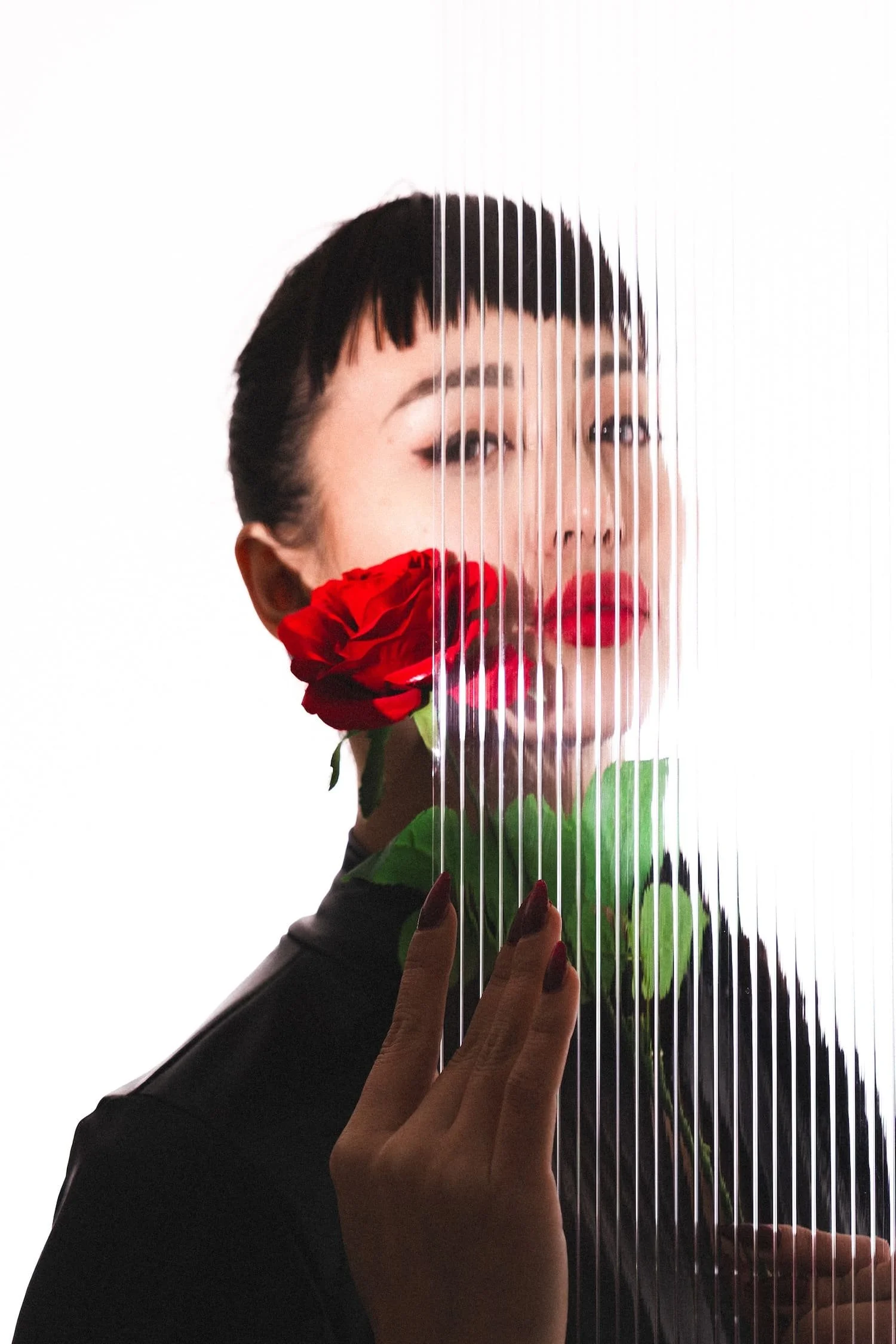 A woman with short black hair and red lipstick holding a red rose behind a transparent textured glass panel, creating a layered artistic portrait.