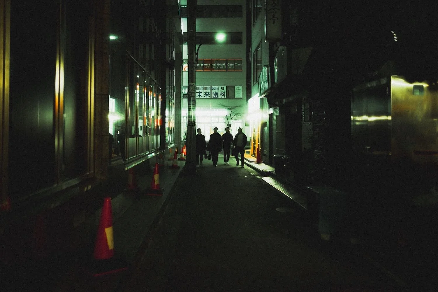Four people walking down a dimly lit city alley at night, with bright green and white lights illuminating the background. Street photography workshop by Deniz Demir Tekumi.