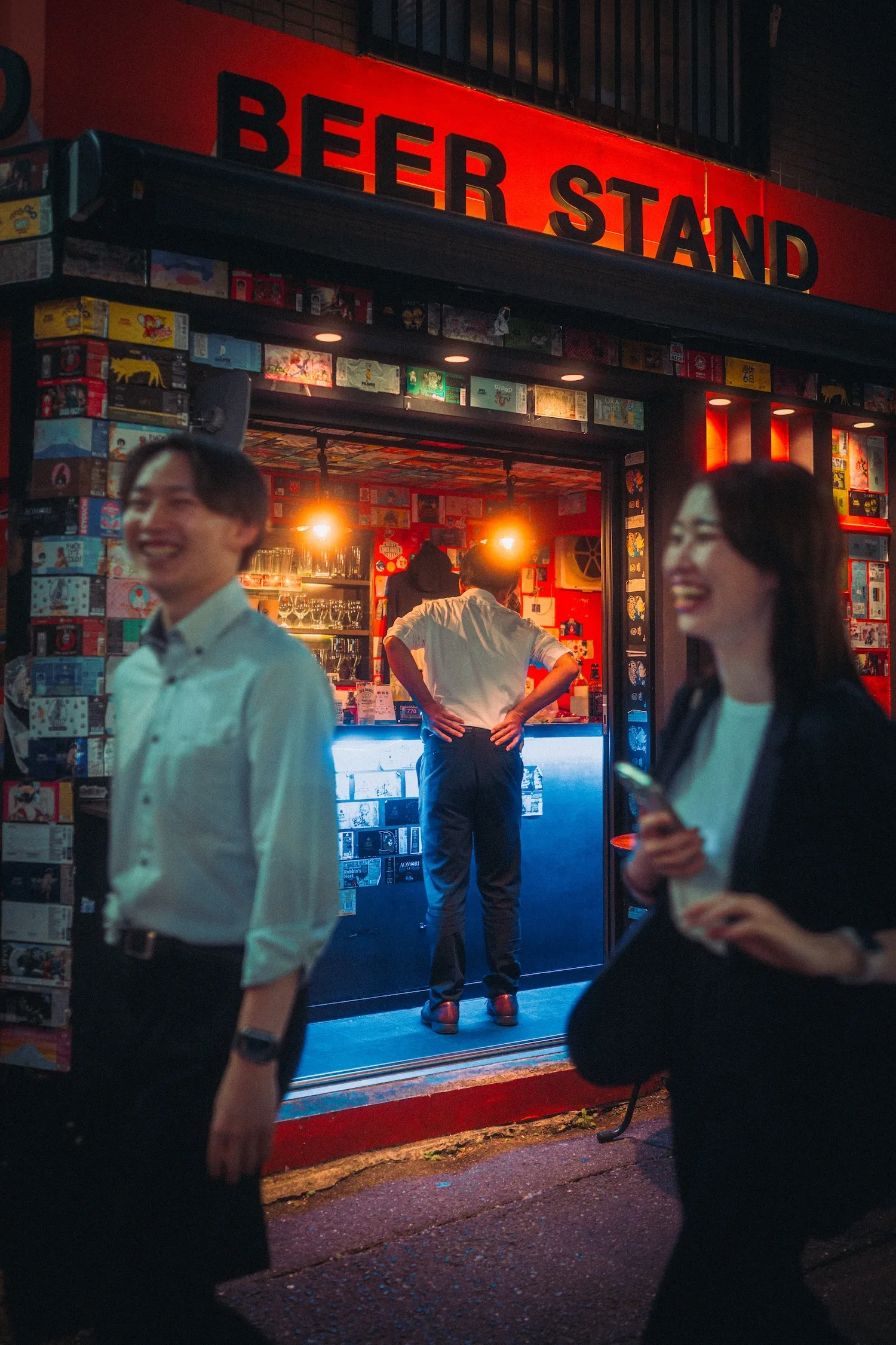 Nighttime scene outside a bar with a large red sign reading "BEER STAND". People are standing and chatting, with some blurred in motion in the foreground. Inside the bar, a person is standing at the counter, and the interior walls are decorated with colorful artwork and posters.