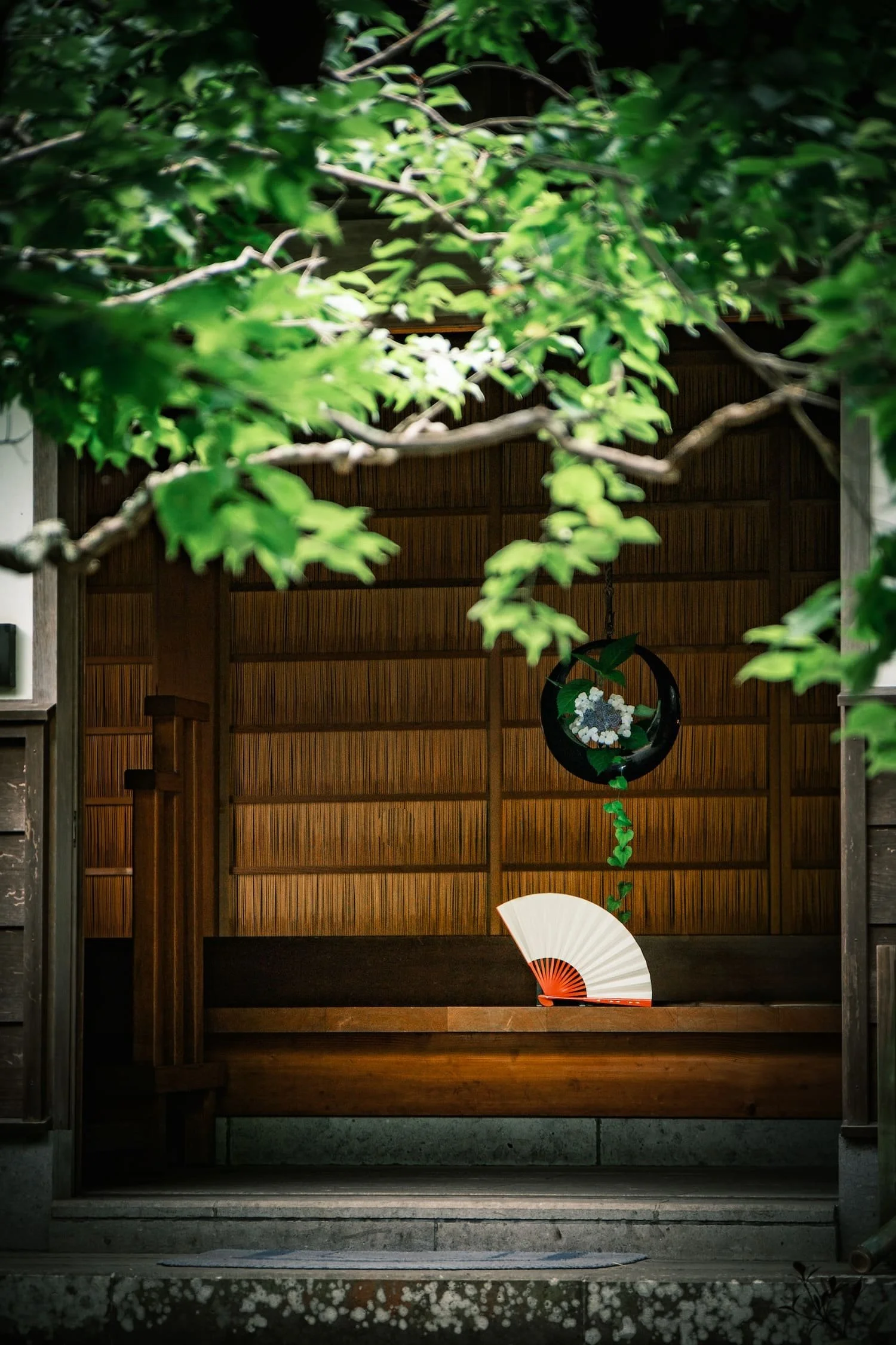 A wooden frame with a roof, featuring a hanging ornament with flowers and a green vine, and a traditional folding fan placed on the ground, partially illuminated.