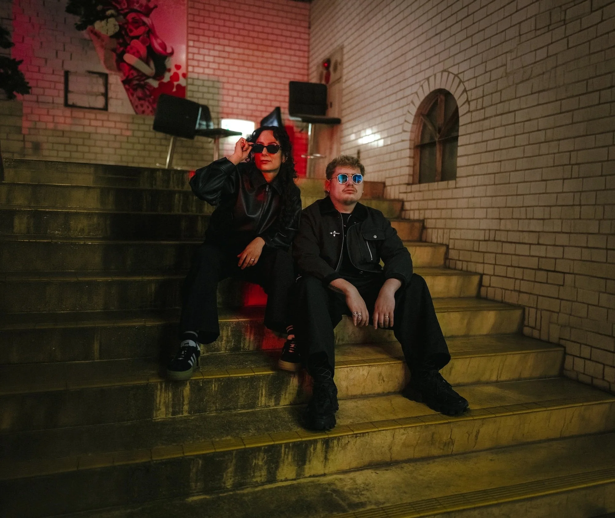 Two people sitting on stairs in an indoor setting with brick walls; the person on the left is wearing dark sunglasses, a black leather jacket, and has curly hair, while the person on the right is wearing reflective sunglasses, a black jacket, and has short hair.