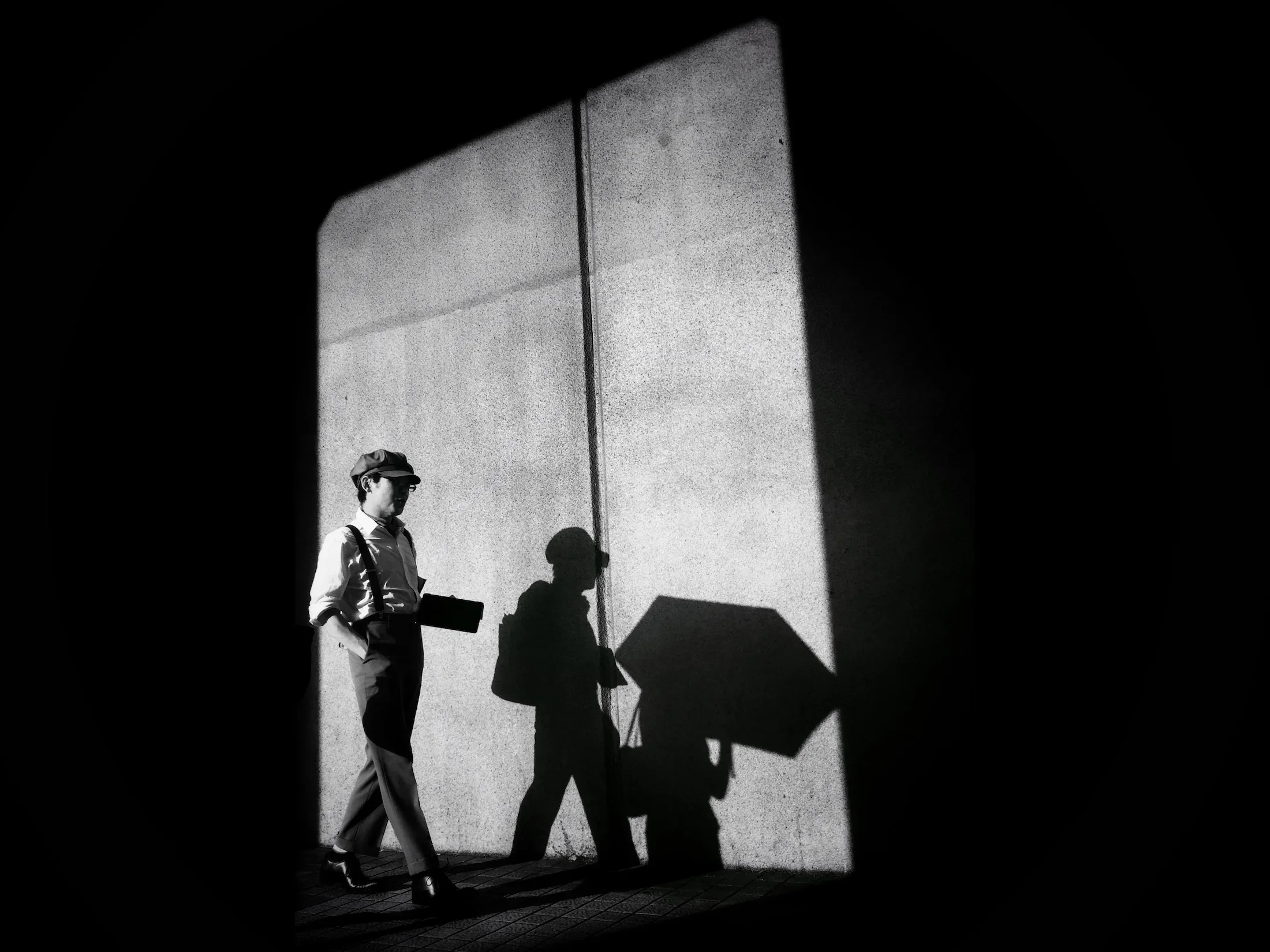A person wearing glasses, a hat, a white shirt, and suspenders walking past a large shadow cast on a concrete wall, with another person's shadow in the background.