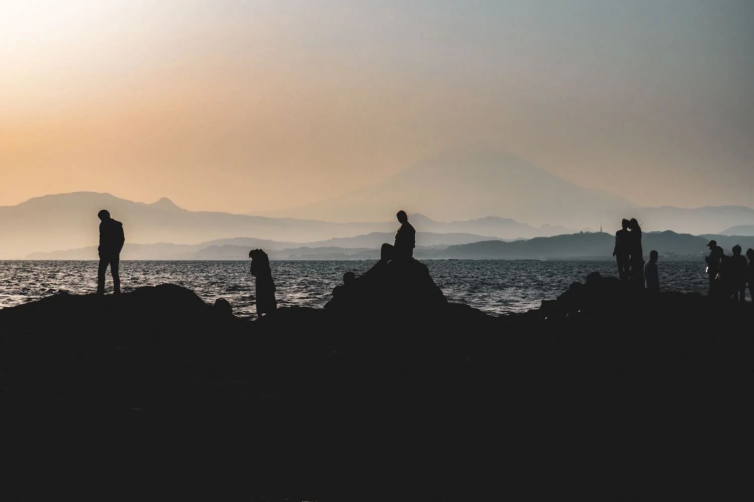 Silhouettes of people sitting and standing on rocks along a coastal area at sunset, with mountains in the background and a calm sea in the foreground.