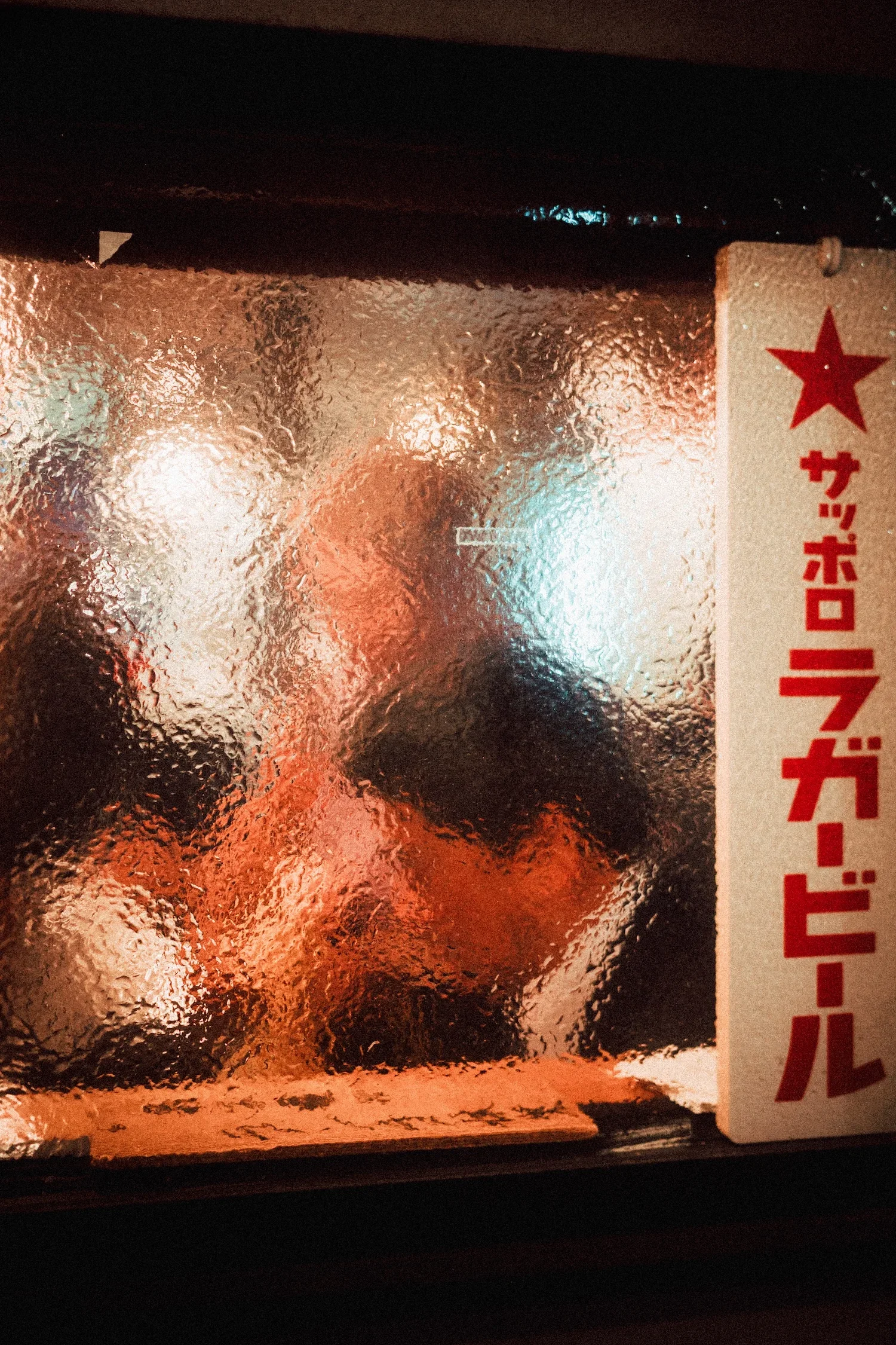A blurry, textured glass window with red and orange lighting behind it. Part of a white sign with red Japanese characters and a red star is visible on the right side of the image.