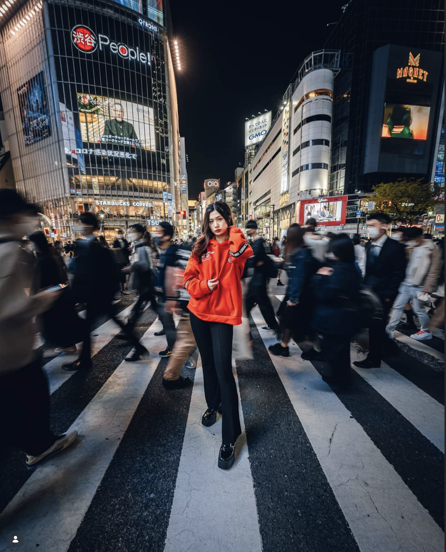A woman in a red hoodie and black pants standing in the middle of a busy city street at night with illuminated buildings and billboards, surrounded by blurred pedestrians.